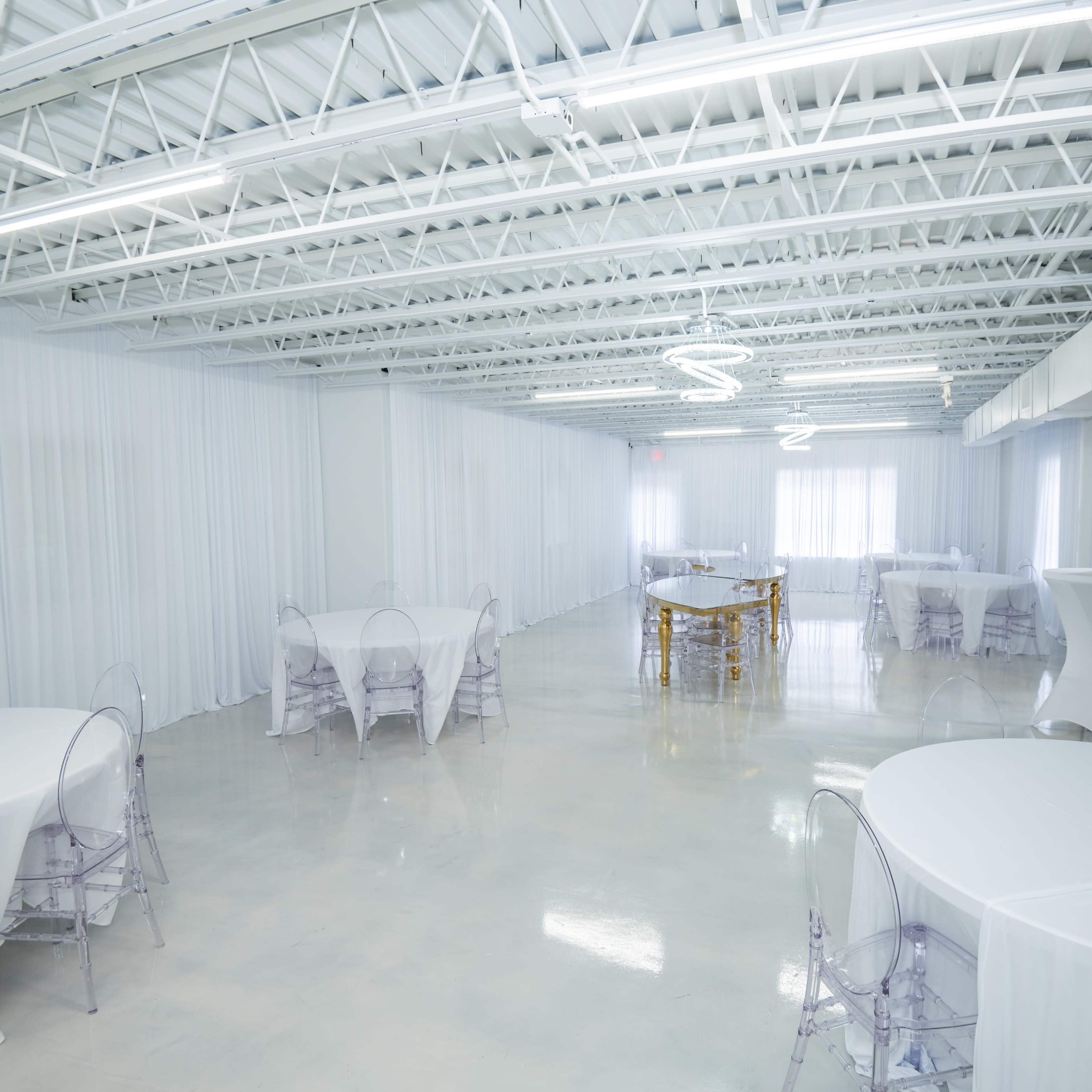 A spacious, well-lit event venue featuring several round tables with white tablecloths and transparent chairs.