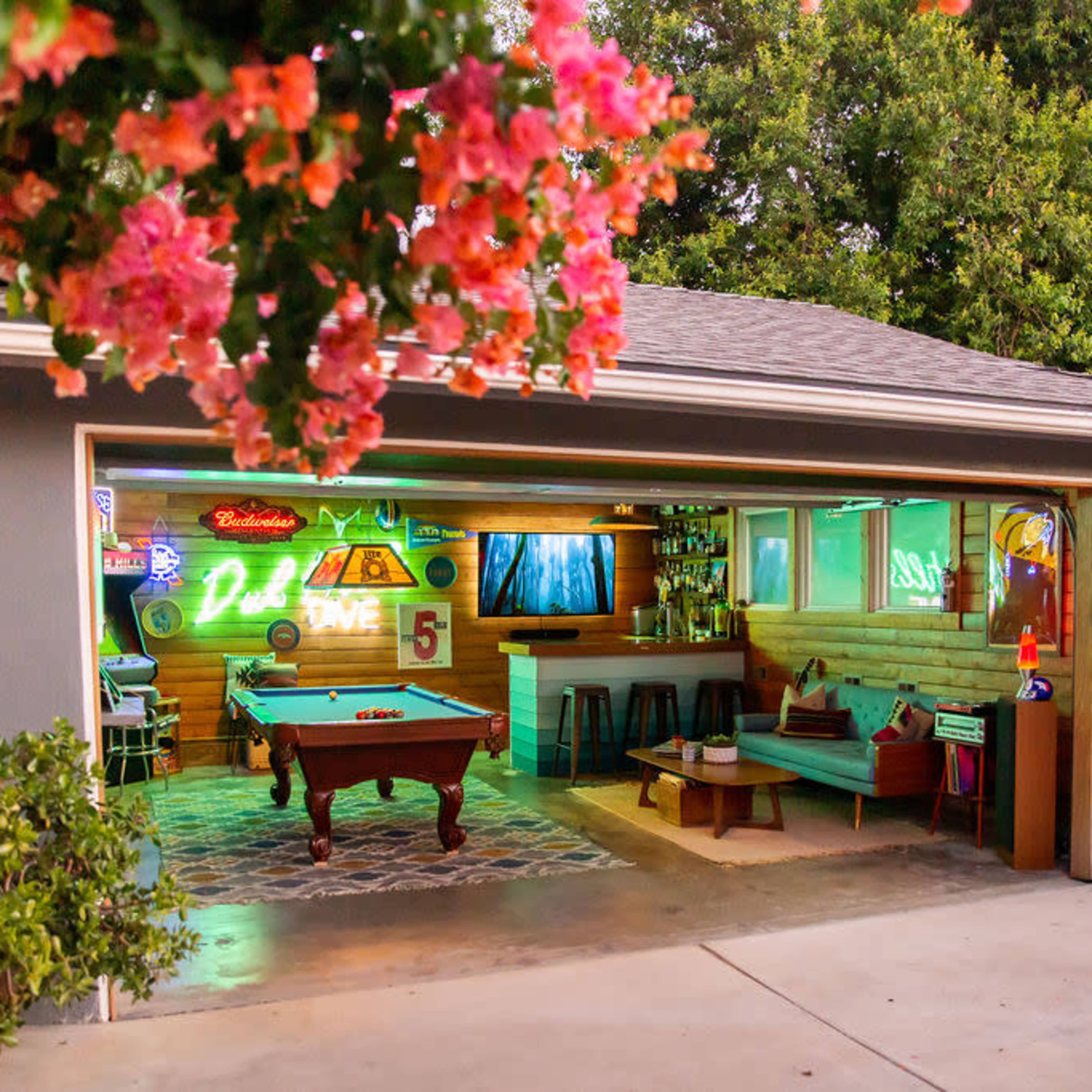 A garage converted into a recreational space with a pool table, neon lights, and a cozy seating area, surrounded by greenery.