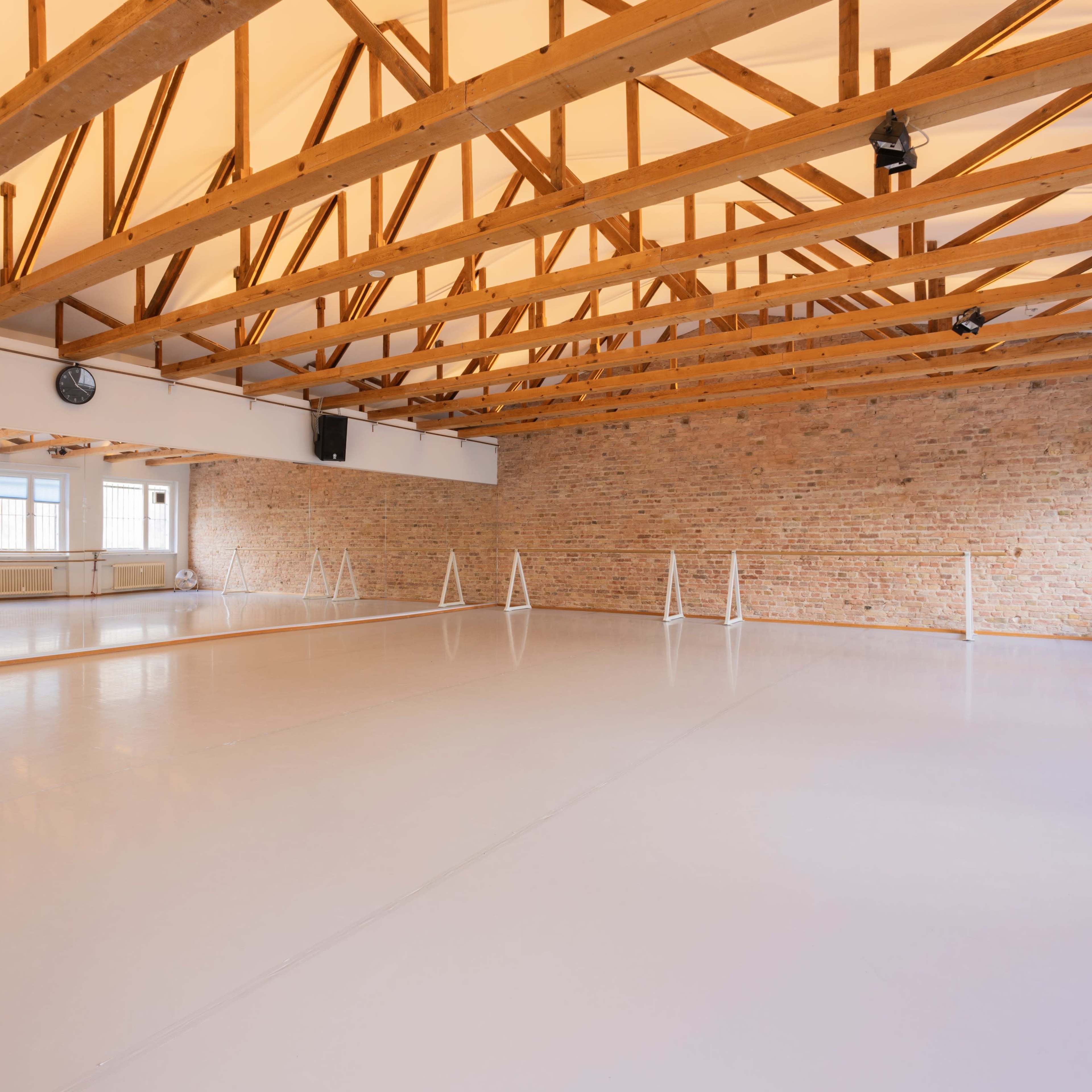A spacious dance studio with wooden beams overhead, large windows allowing natural light, and a smooth floor with ballet barres along the walls.