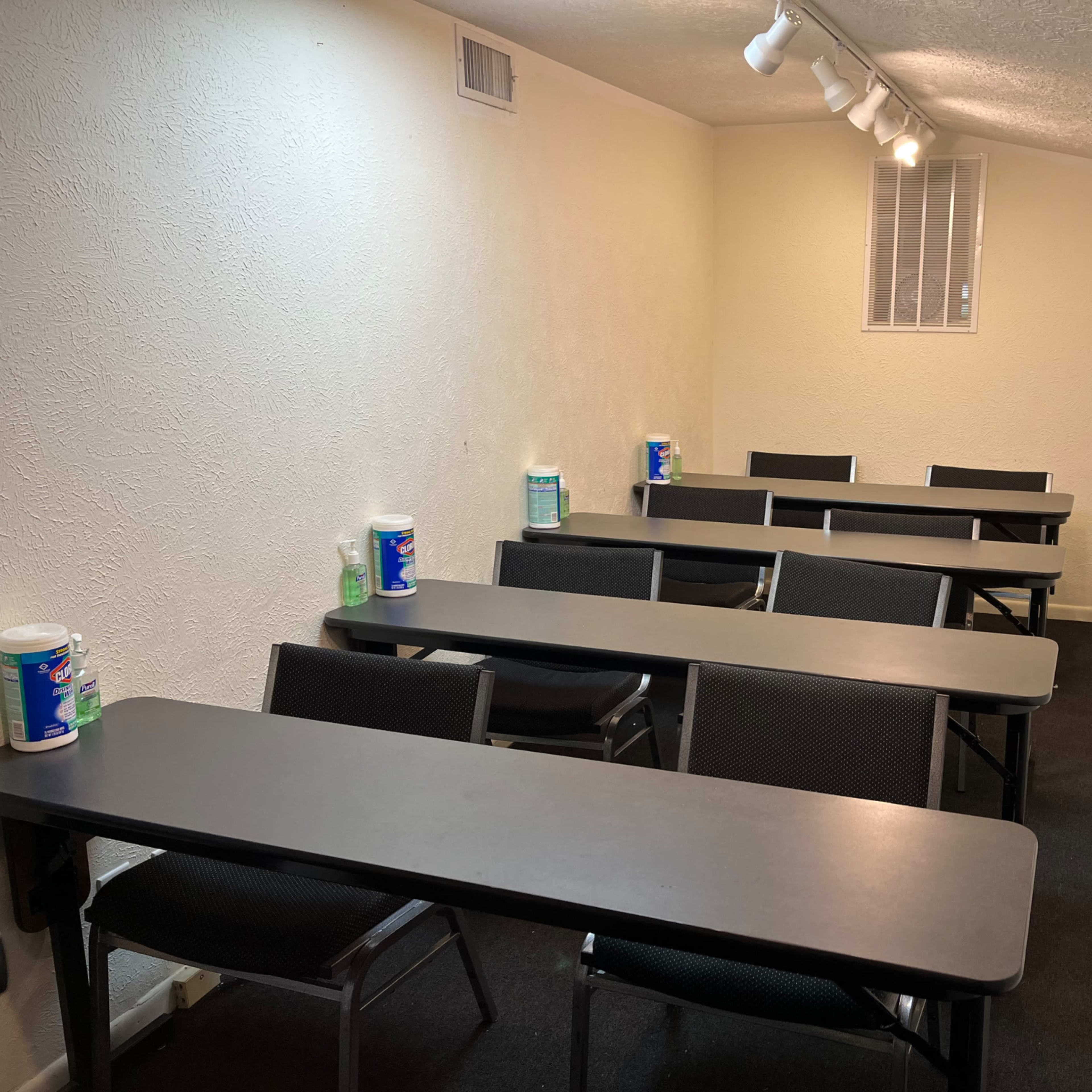 A narrow classroom features several rows of black tables and chairs positioned on a dark carpeted floor, with disinfectant supplies placed on the tables.