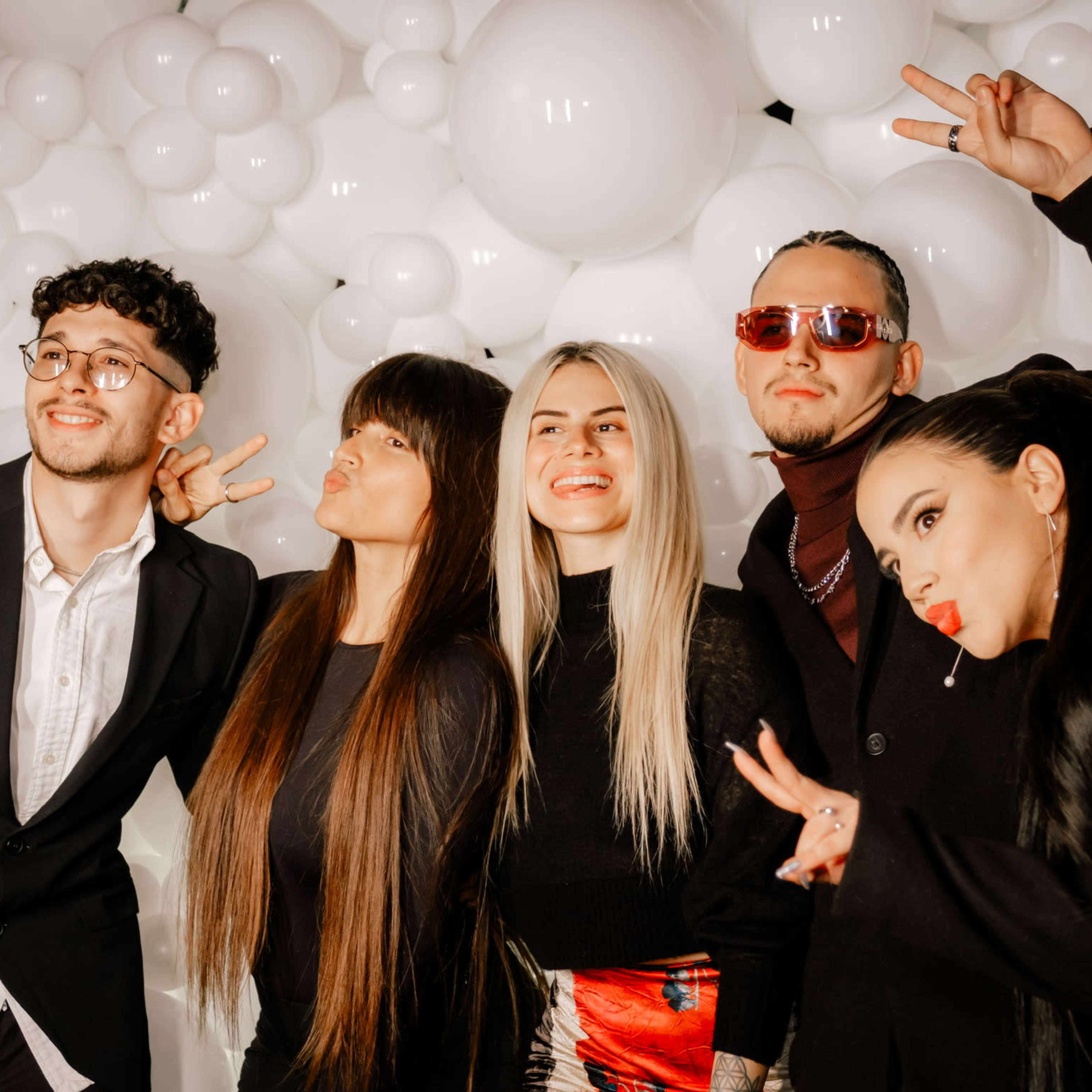 A group of five individuals poses with peace signs in front of a backdrop of large white balloons.