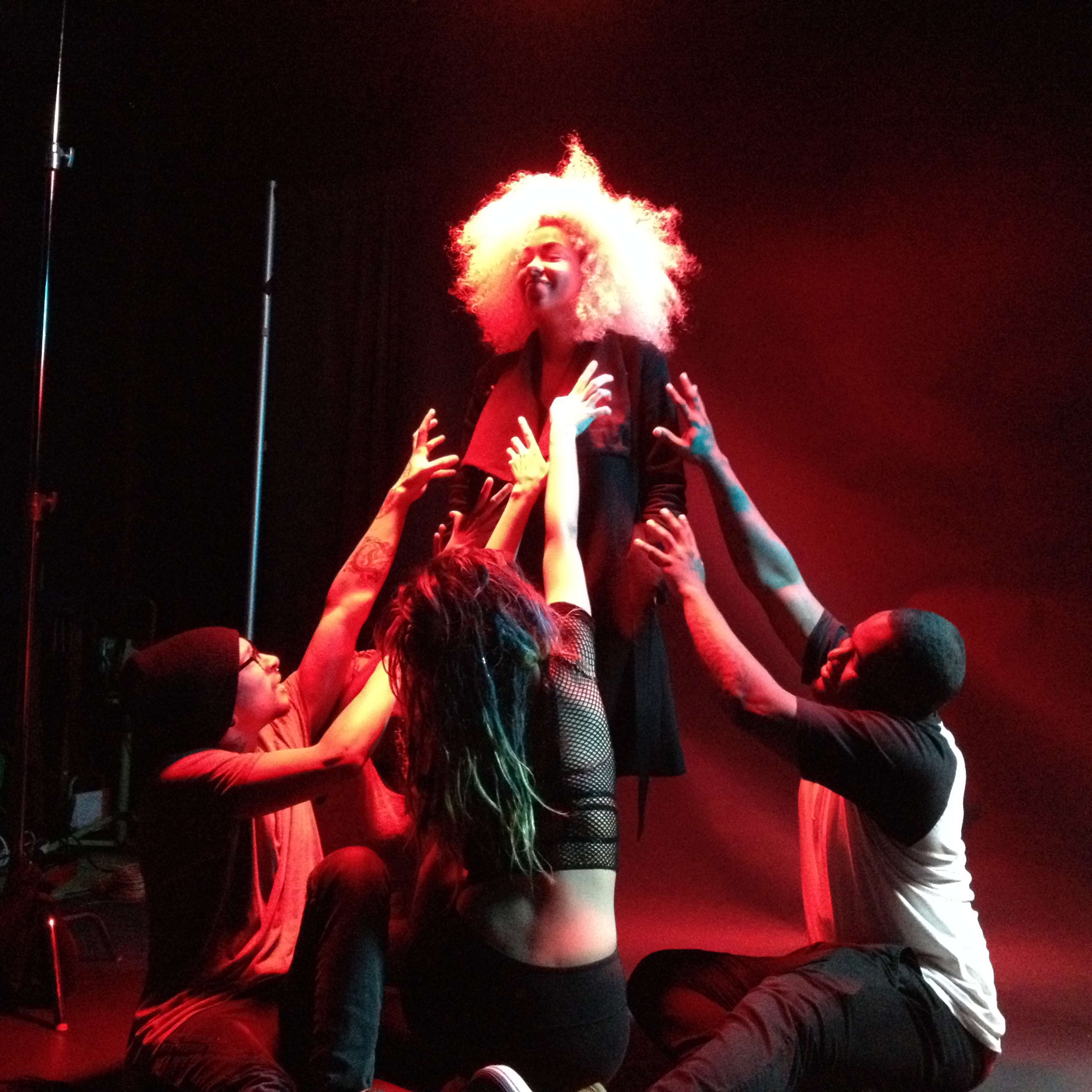 A person with an elaborate hairstyle is surrounded by four individuals, who are reaching towards them on a stage with dramatic lighting.