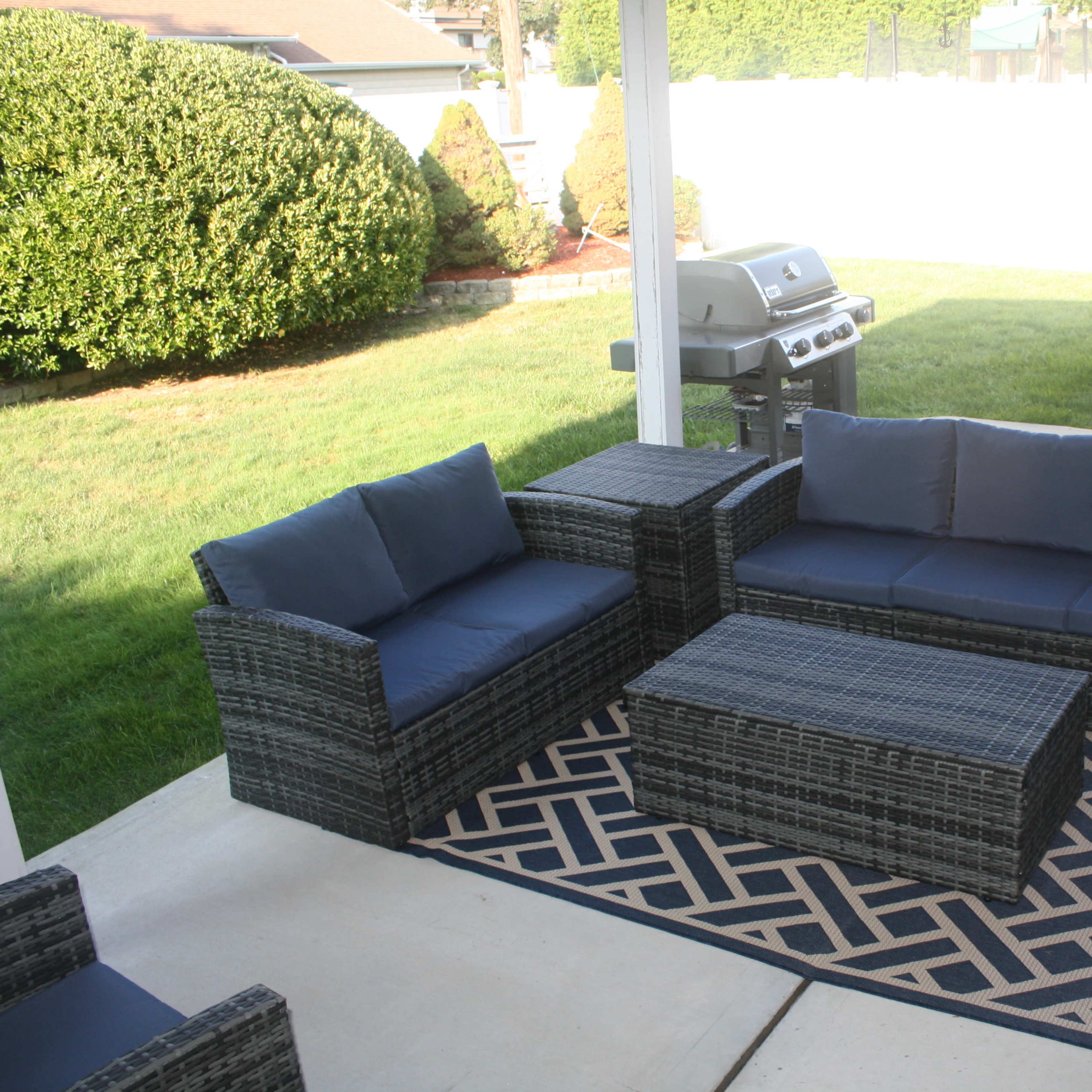 A patio area with outdoor furniture, including a sofa set, coffee table, and a gas grill, surrounded by a green lawn and a hot tub.