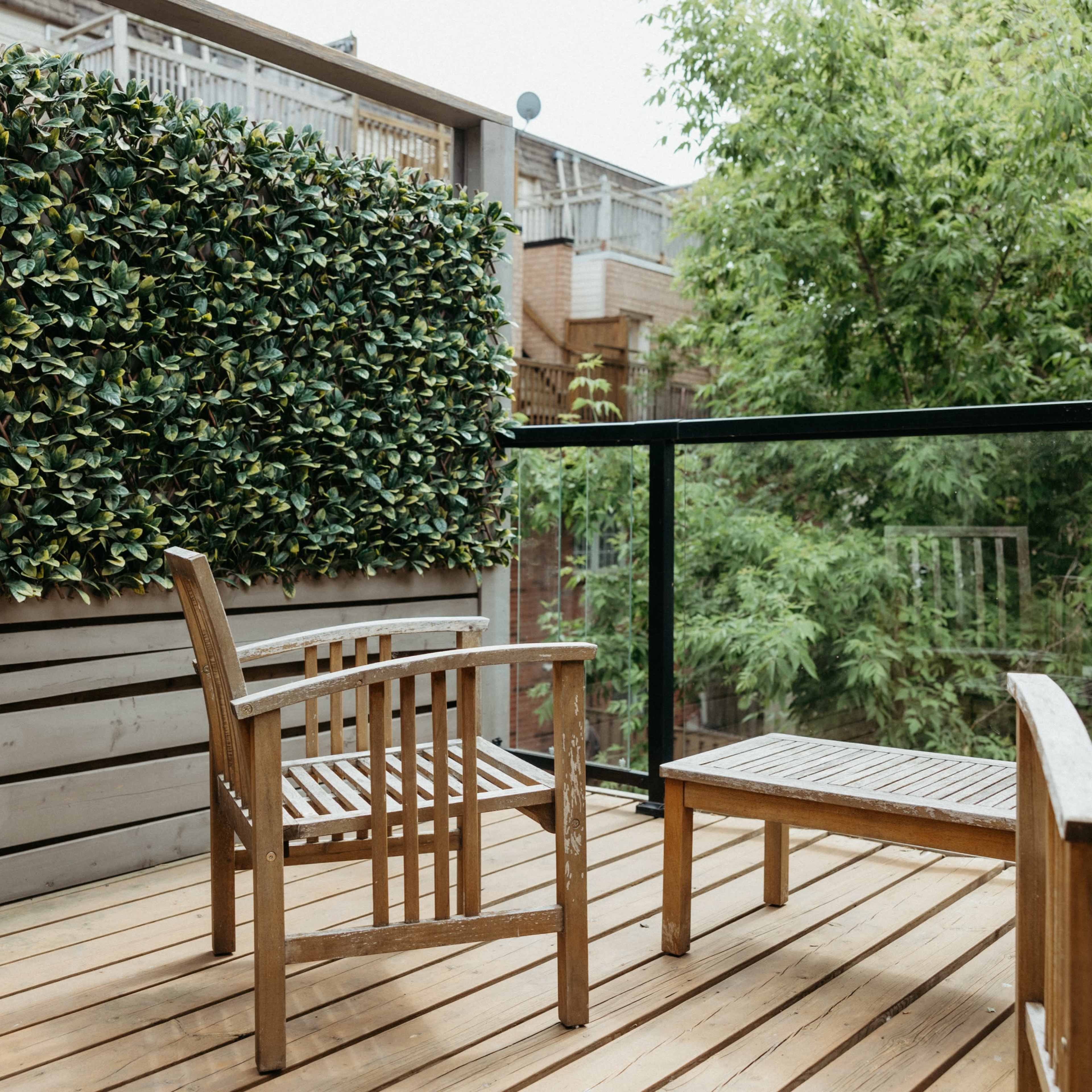 A backyard deck features two wooden chairs and a small table, surrounded by greenery and a privacy screen.