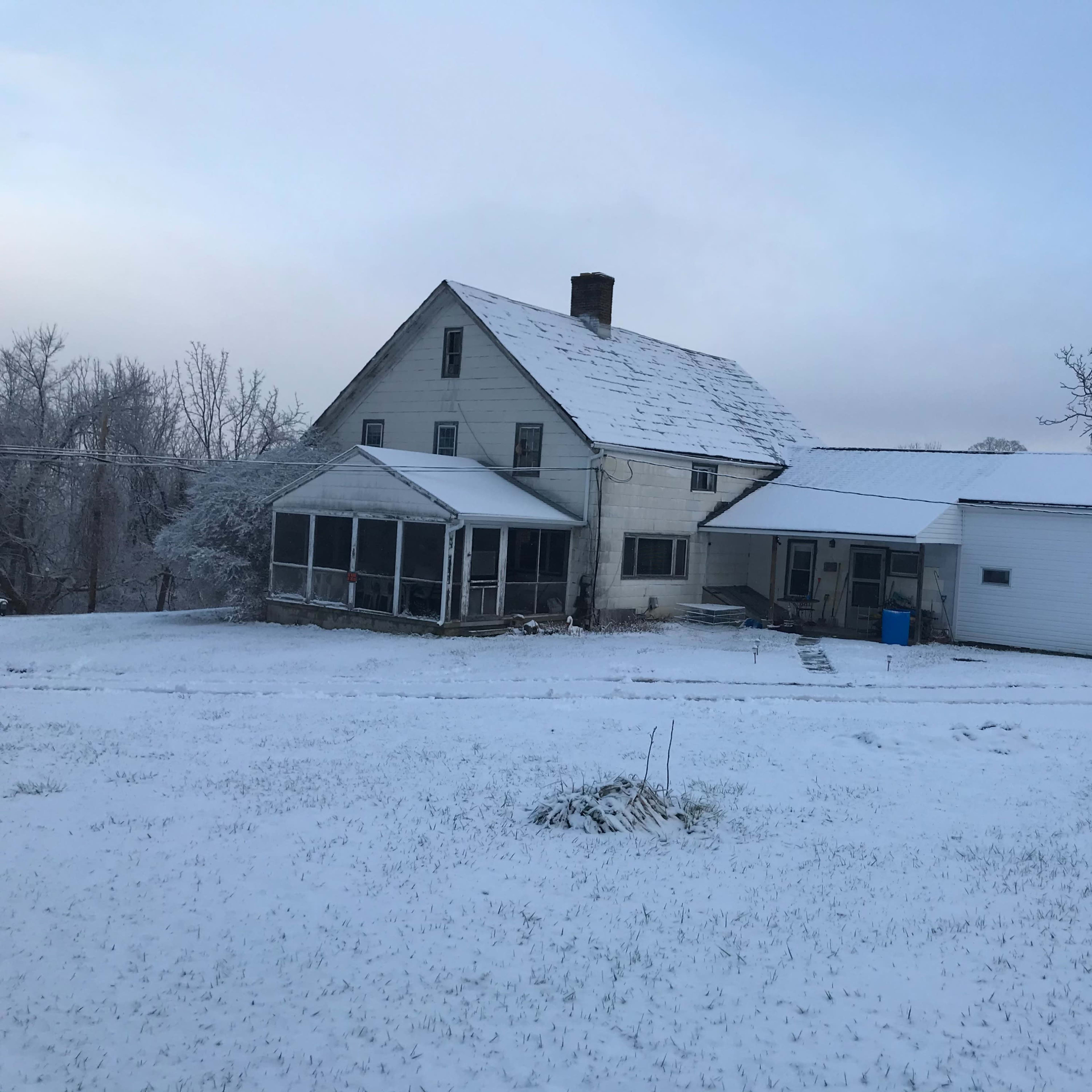 A weathered house with a screened porch and an attached garage is surrounded by a blanket of snow.