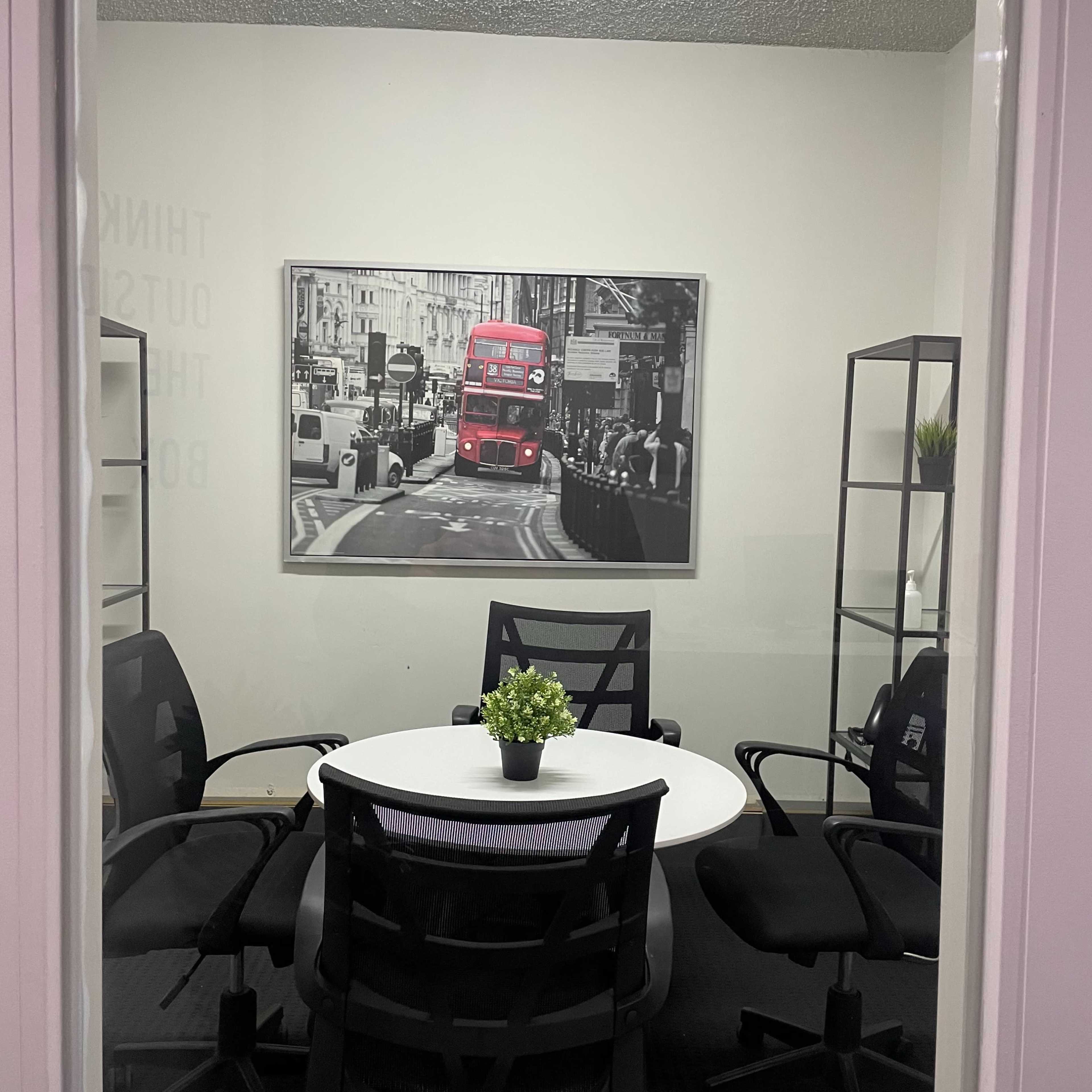 A round table with black chairs is set up in a small conference room, featuring a framed black-and-white photo of a red double-decker bus on the wall.