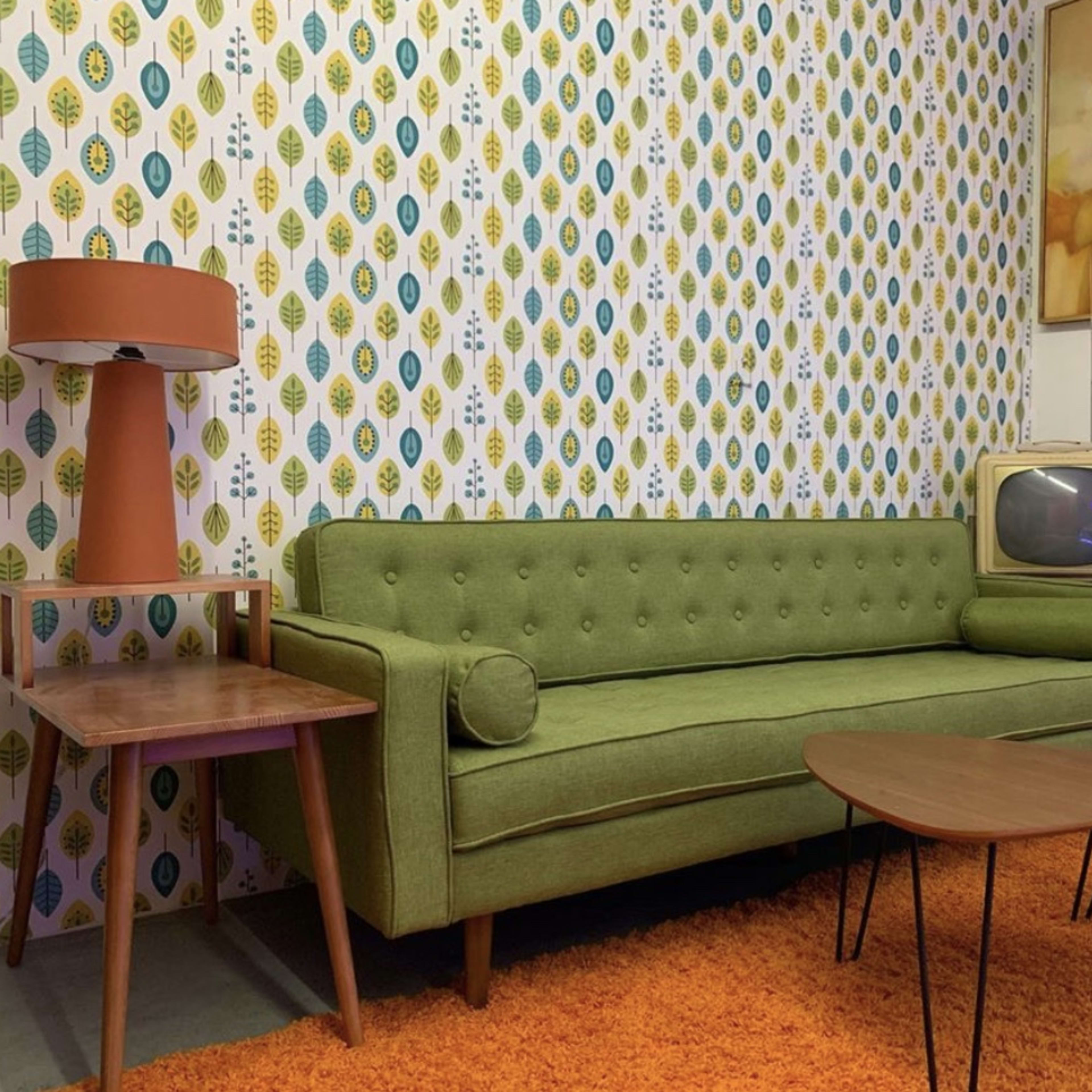 A vintage living room with a green sofa, a side table, a retro lamp, and a small television set against a patterned wallpaper.