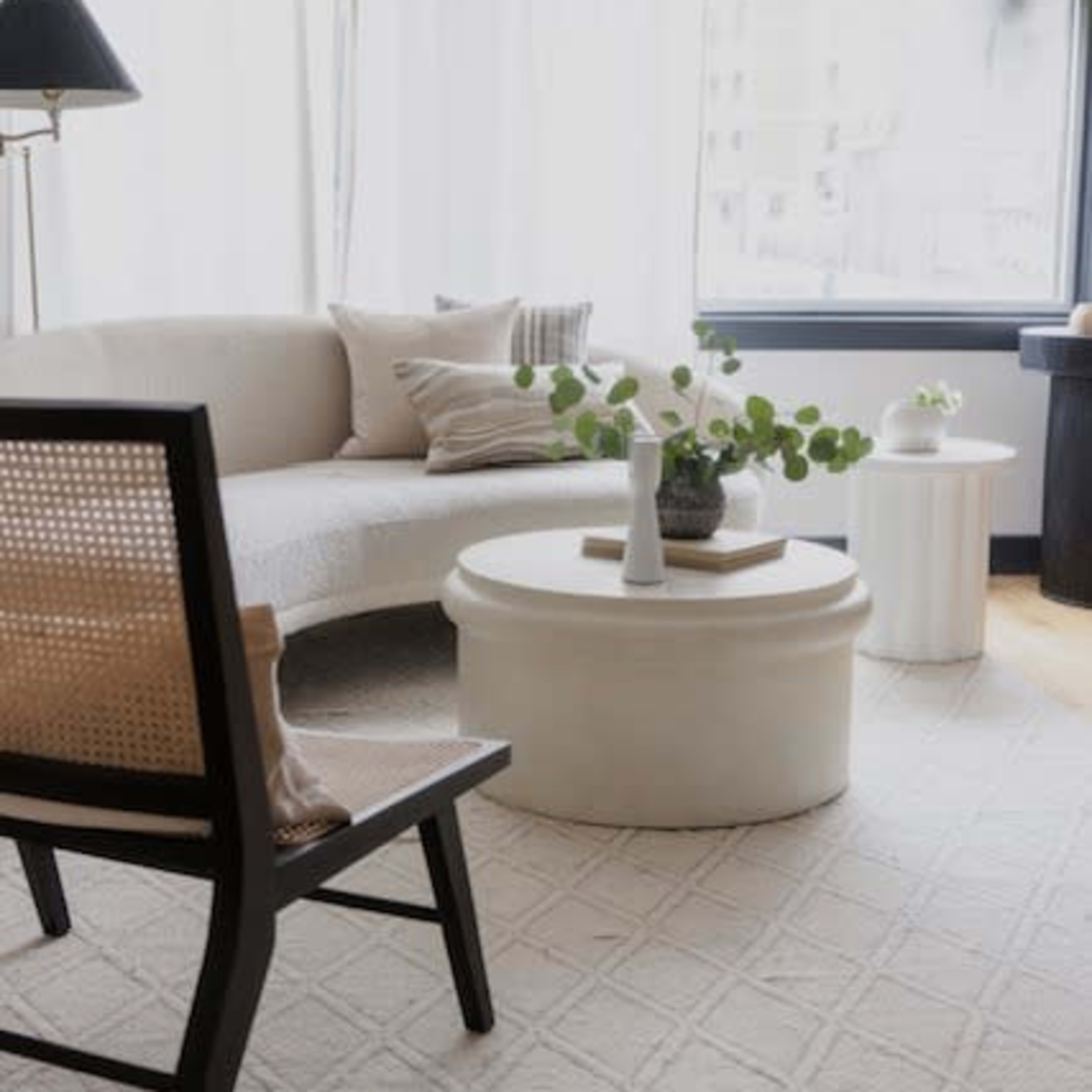 A modern living room with a curved sofa, a round coffee table, and a chair, all set against large windows draped with sheer curtains.