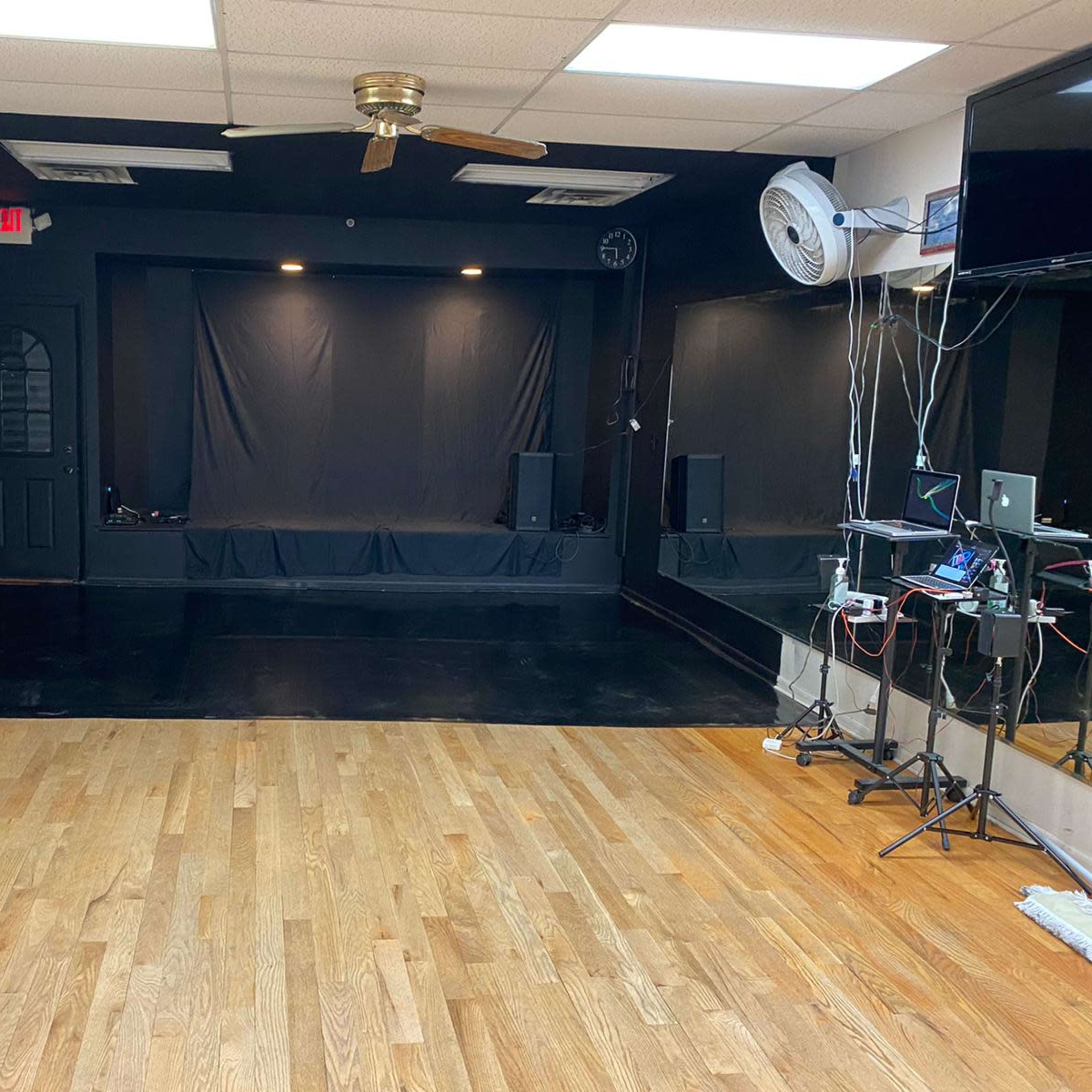 A spacious empty performance room with black curtains, a wooden floor, and music equipment set up on one side.