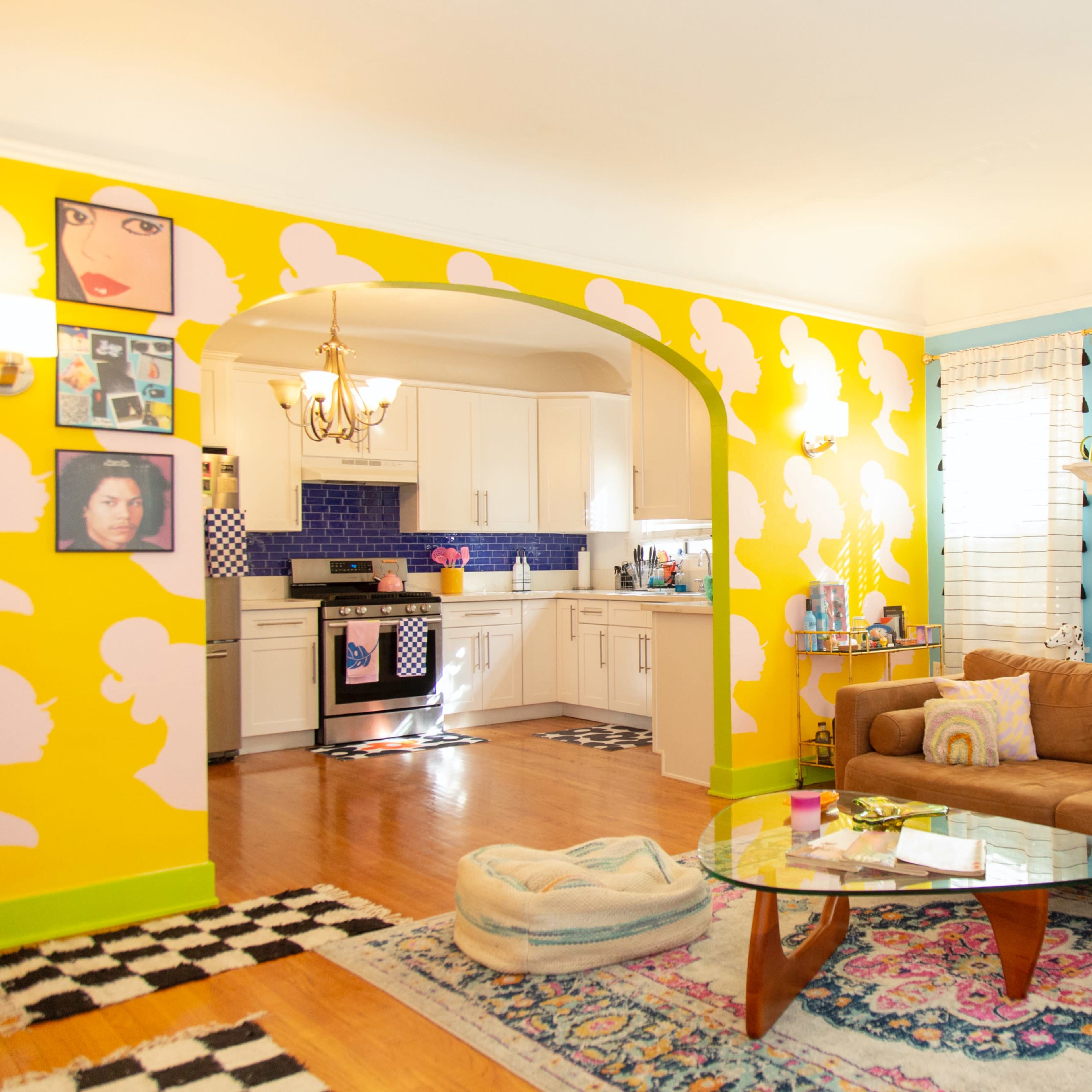 The living room features a brightly painted yellow wall with cloud patterns, a cozy brown sofa, a glass coffee table, and a white fireplace surrounded by colorful artwork.