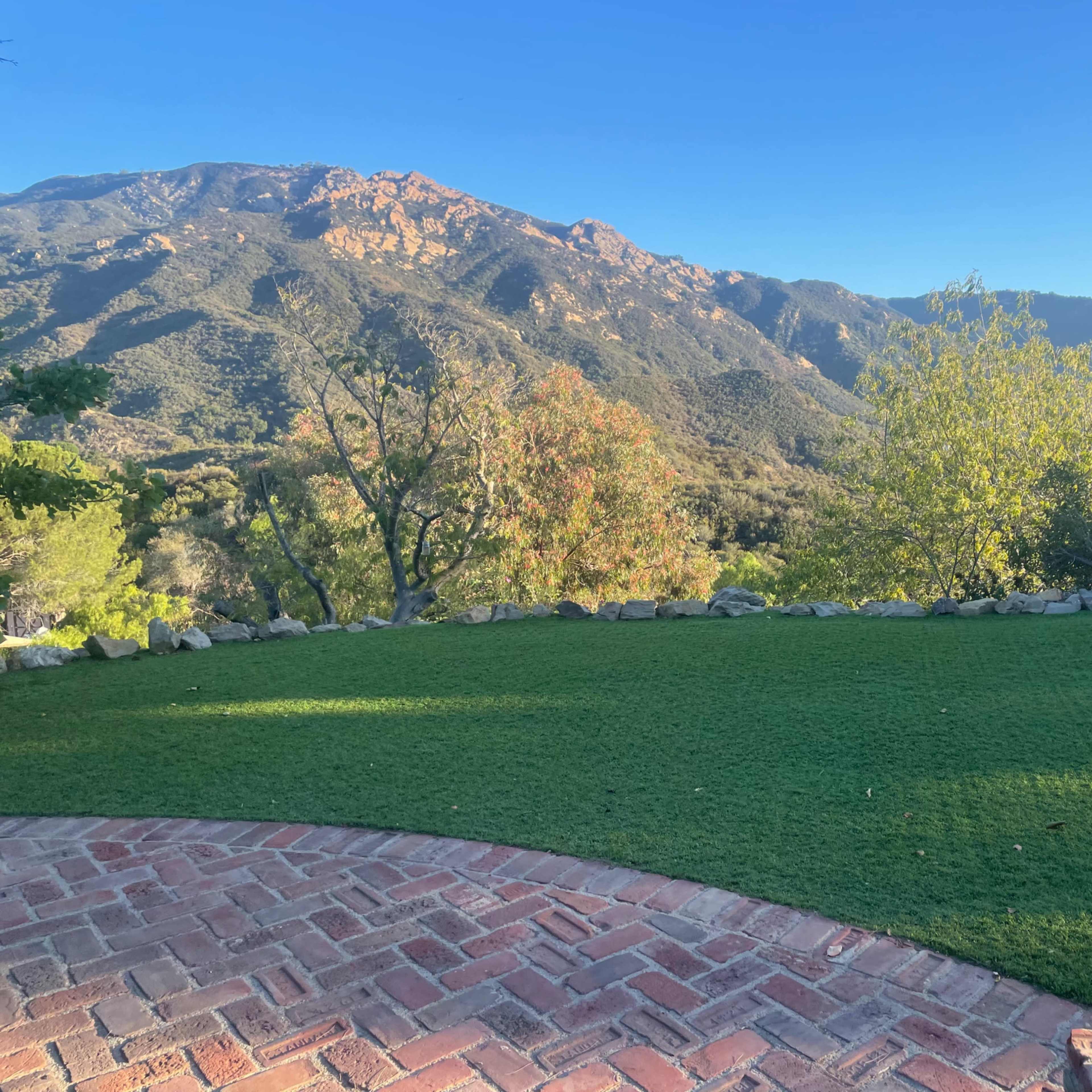 A view of a grassy area bordered by a stone wall, overlooking a mountainous landscape under a clear blue sky.