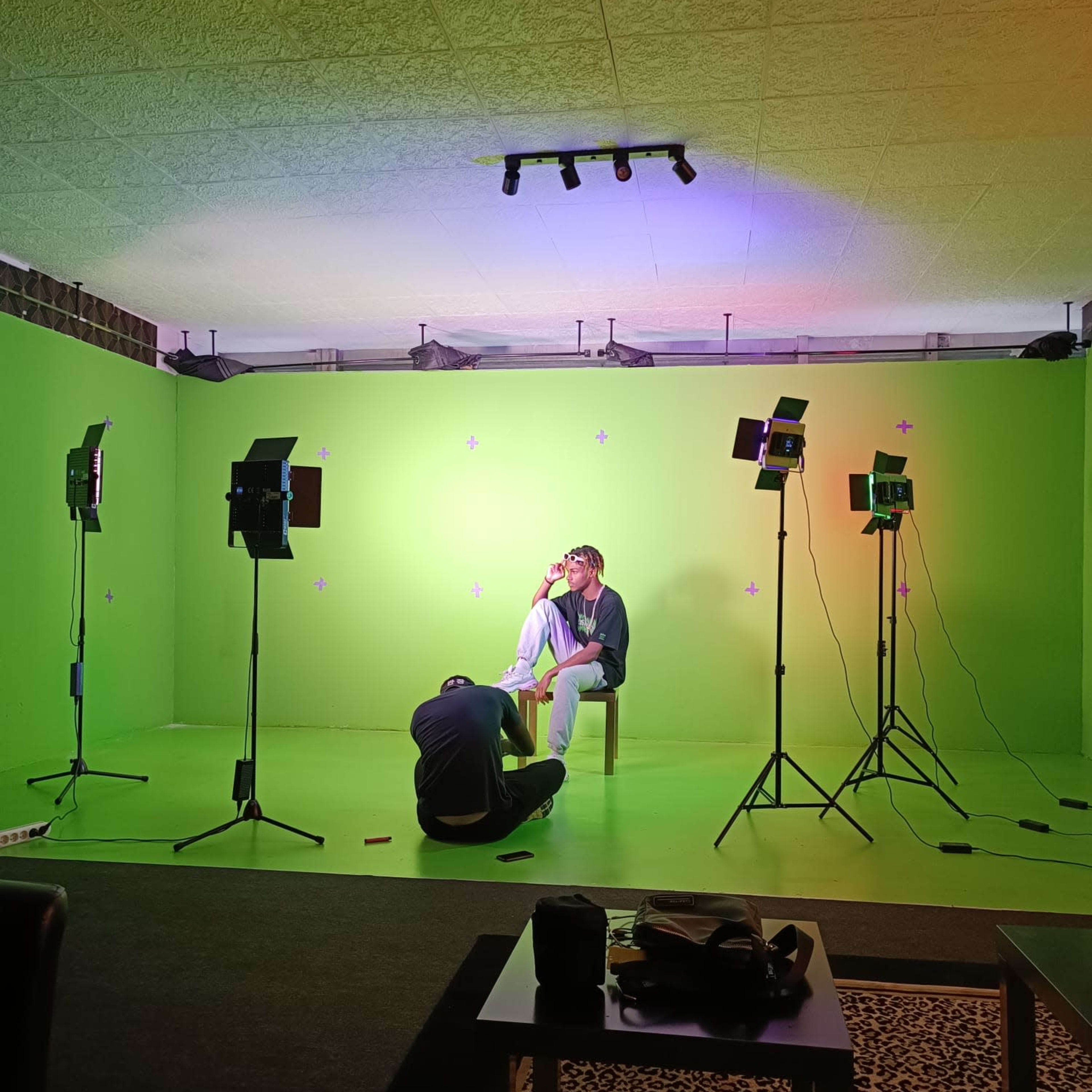 A person sits on a chair in a brightly colored green room surrounded by several studio lights and another individual on the floor nearby.
