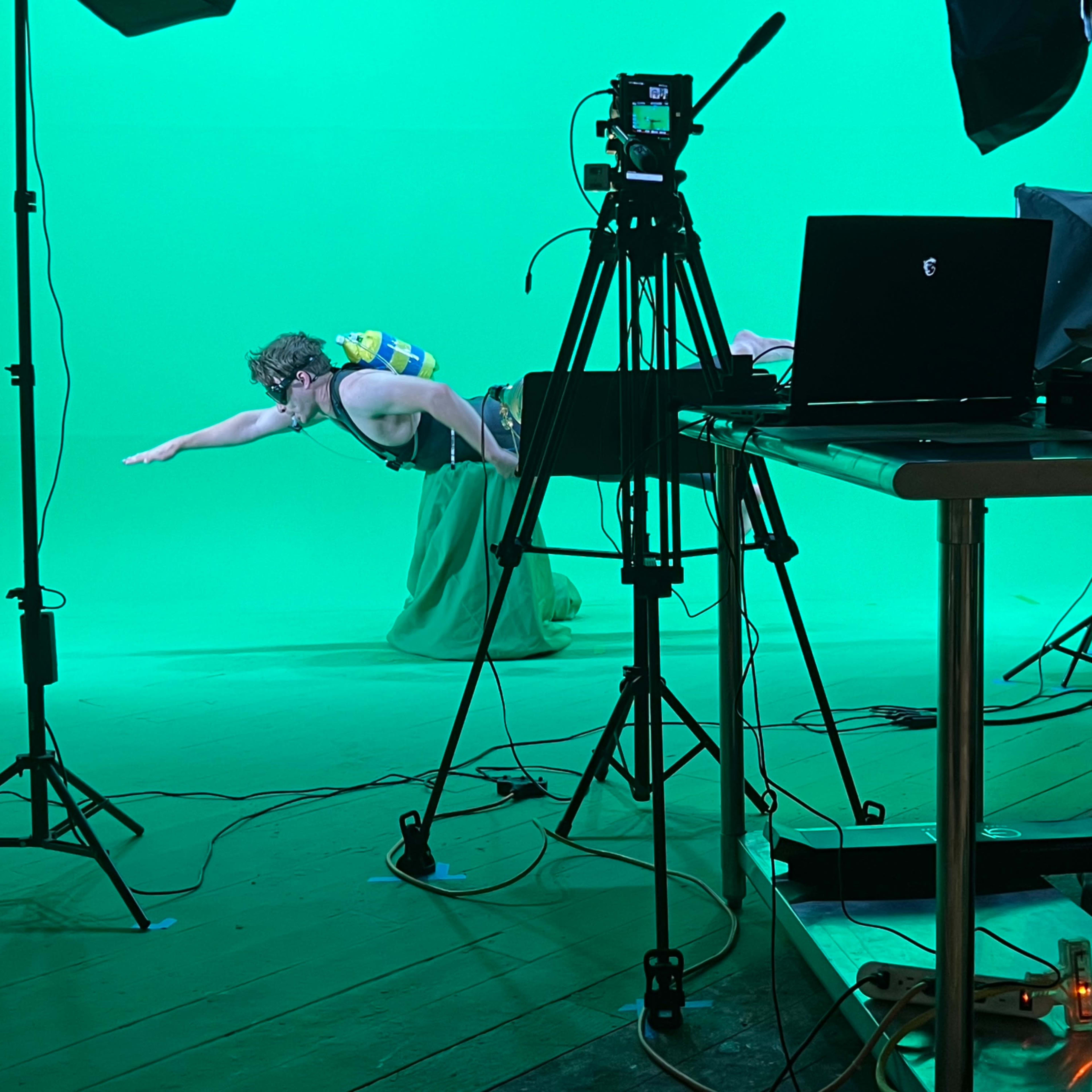 A person is swimming in an indoor studio with a green screen backdrop, surrounded by camera equipment and a laptop on a table.