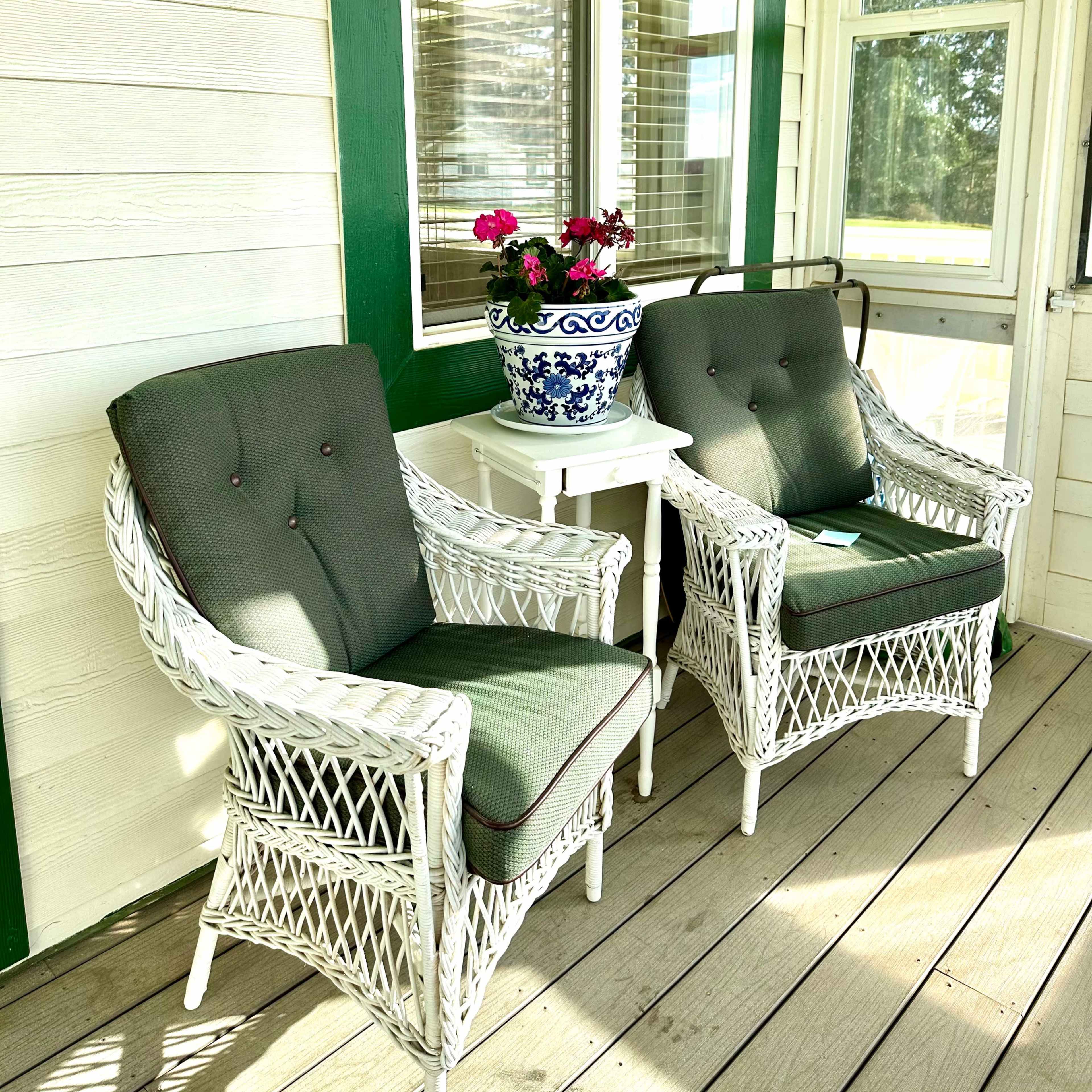Two white wicker chairs with green cushions positioned on a porch beside a table and a flower pot.