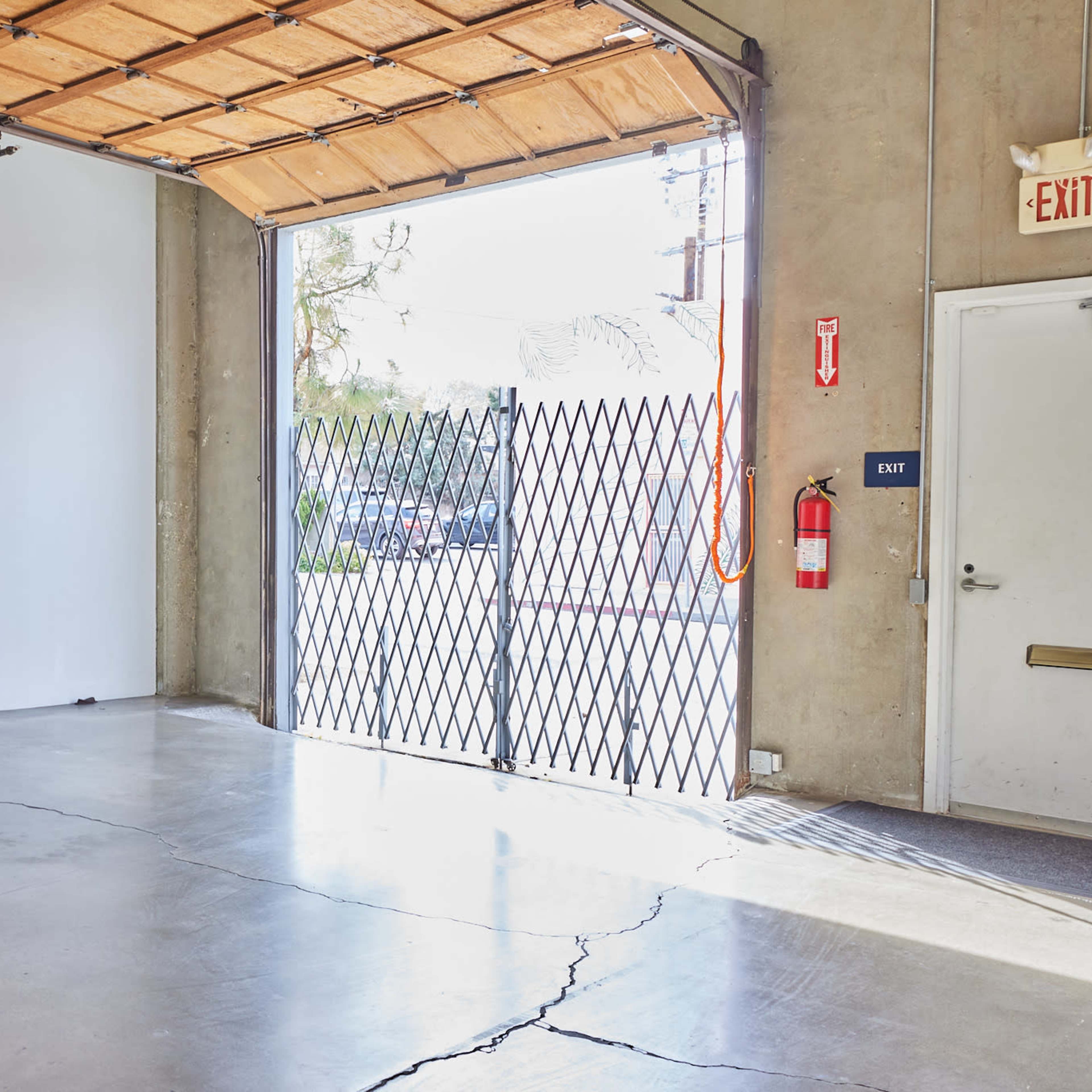 A partially open garage door leading to an outdoor area, with a concrete floor and a visible crack in the surface.