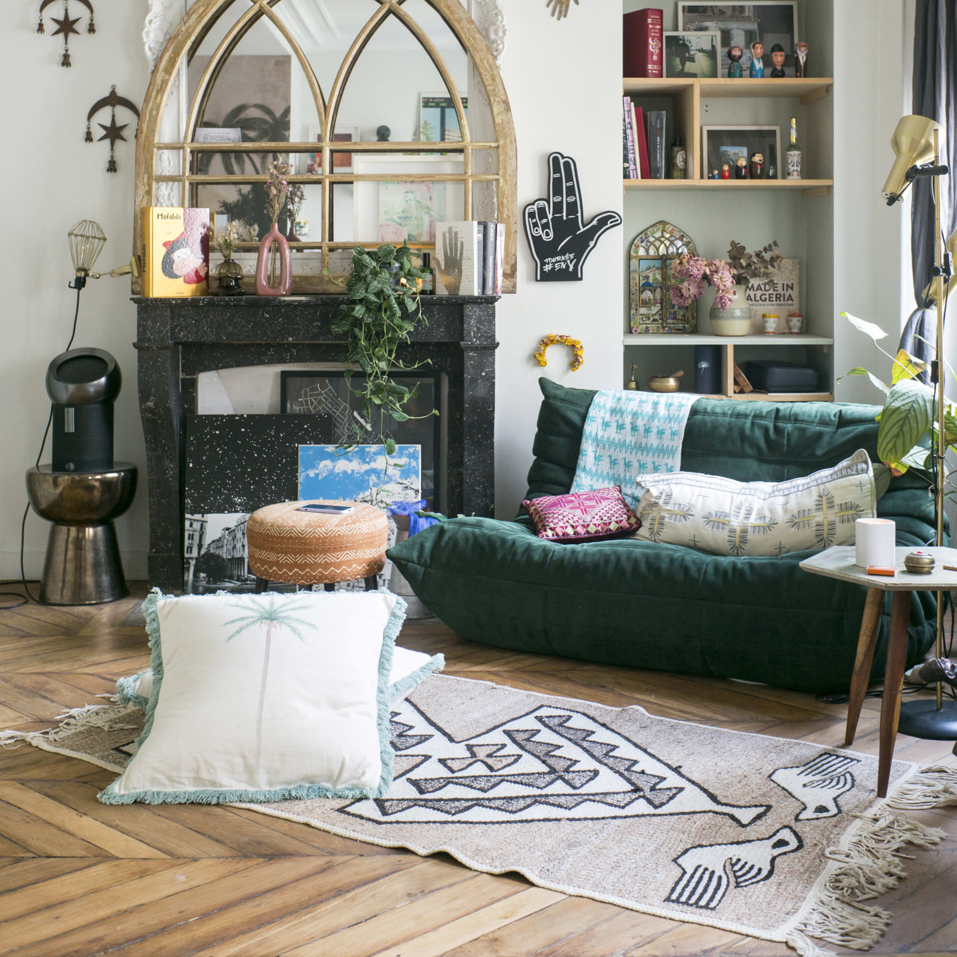 A cozy living room features a dark green sofa, a large arched mirror above a black fireplace, and a patterned rug on a wooden floor, accented by decorative shelves and plants.