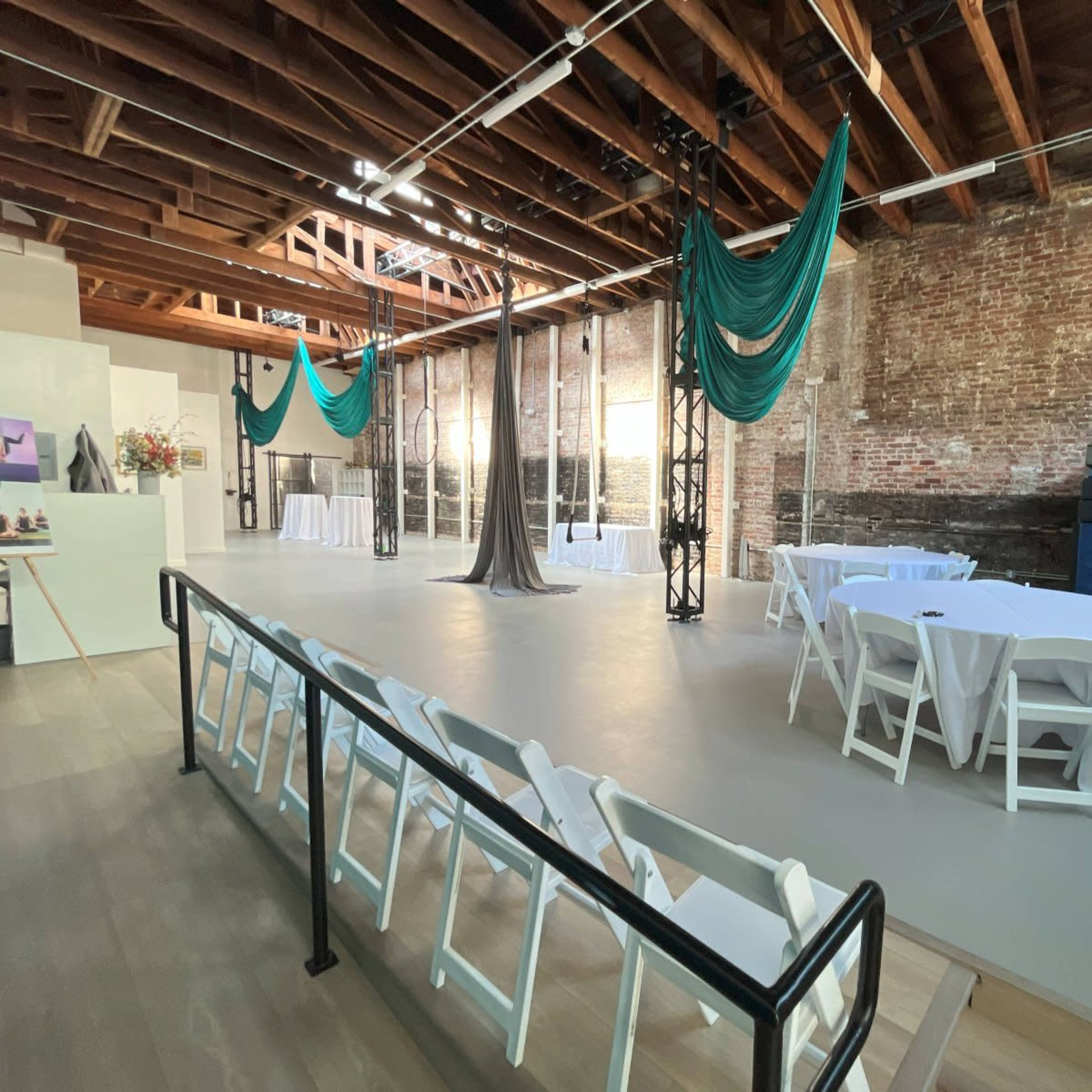 A spacious venue with exposed brick walls, white tables set with tablecloths, and teal fabric draped from the ceiling.