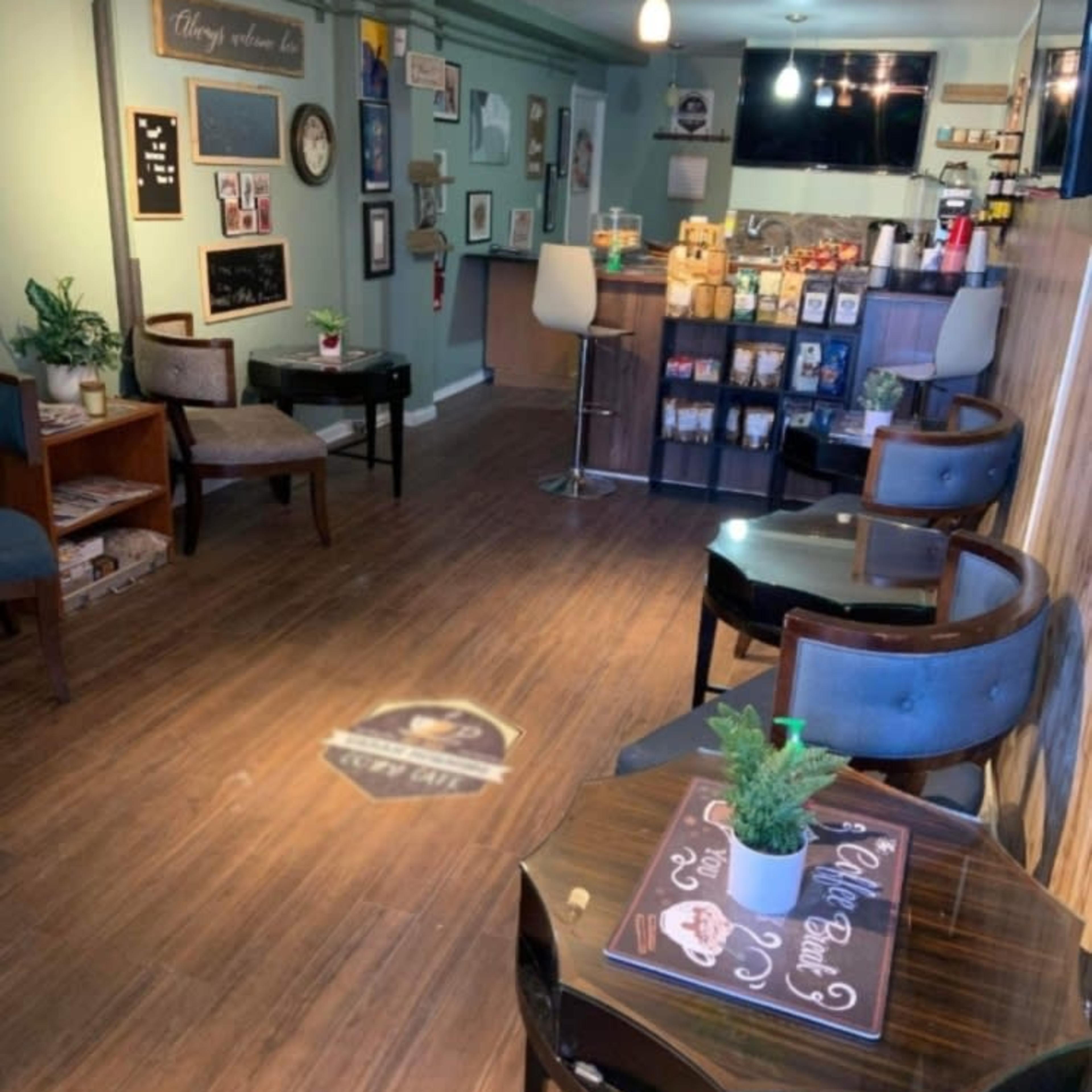 A cozy café interior with wooden flooring, a small seating area with chairs and tables, and a counter displaying various food and beverage items.