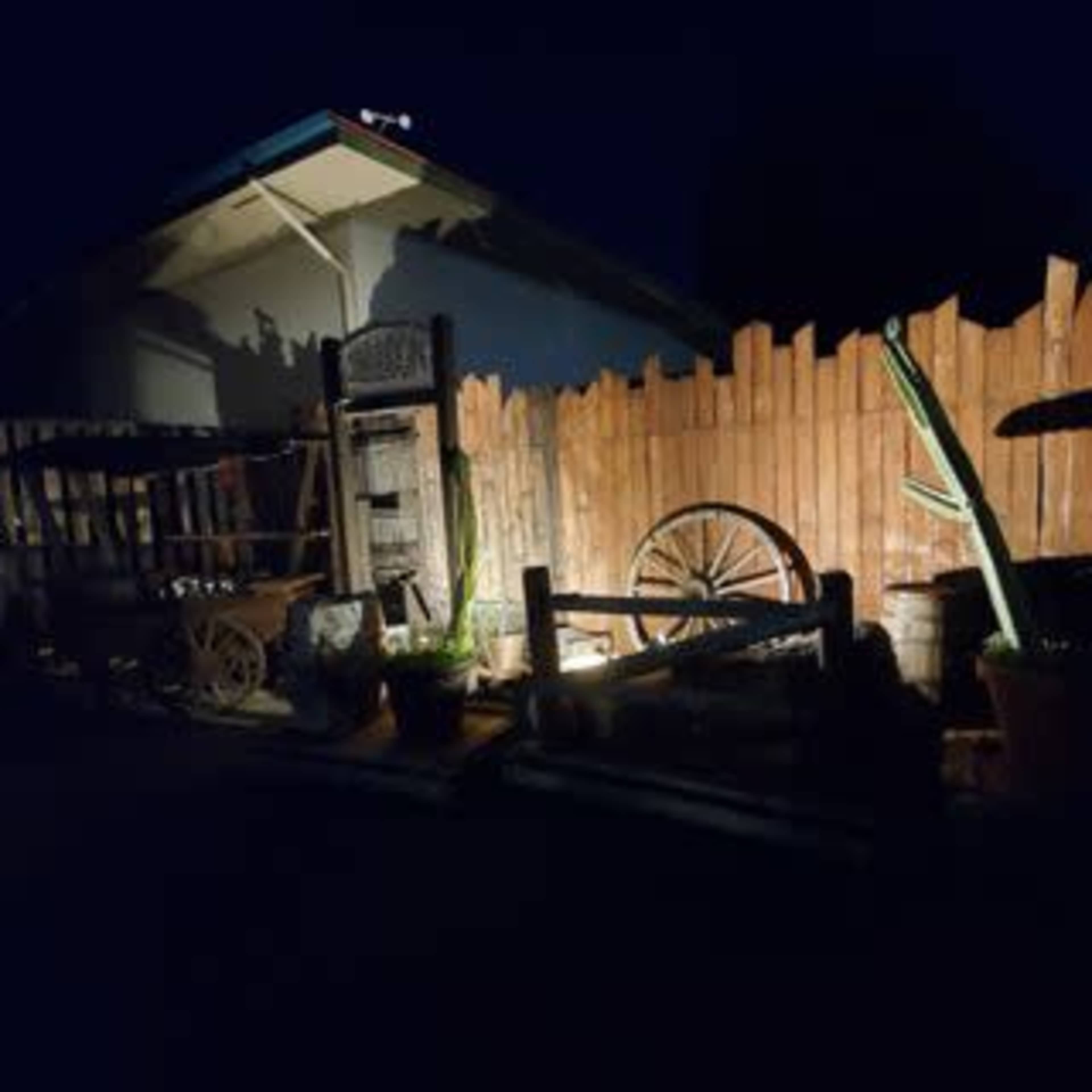 A dimly lit outdoor scene featuring wooden fences, a cartwheel, pots, and a cactus against a house at night.