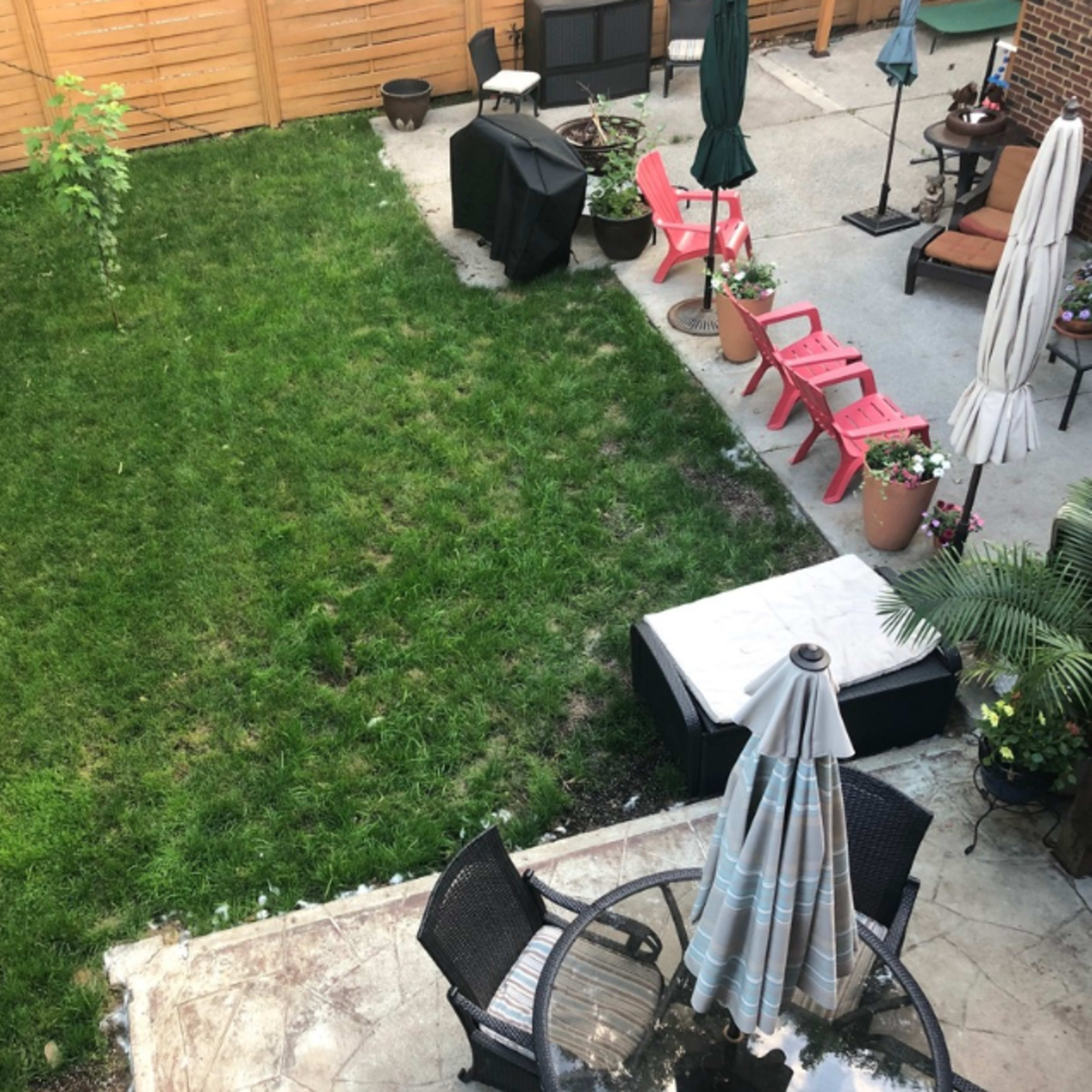 A backyard with a small lawn, a patio area with chairs and a table, a grill, and potted plants.