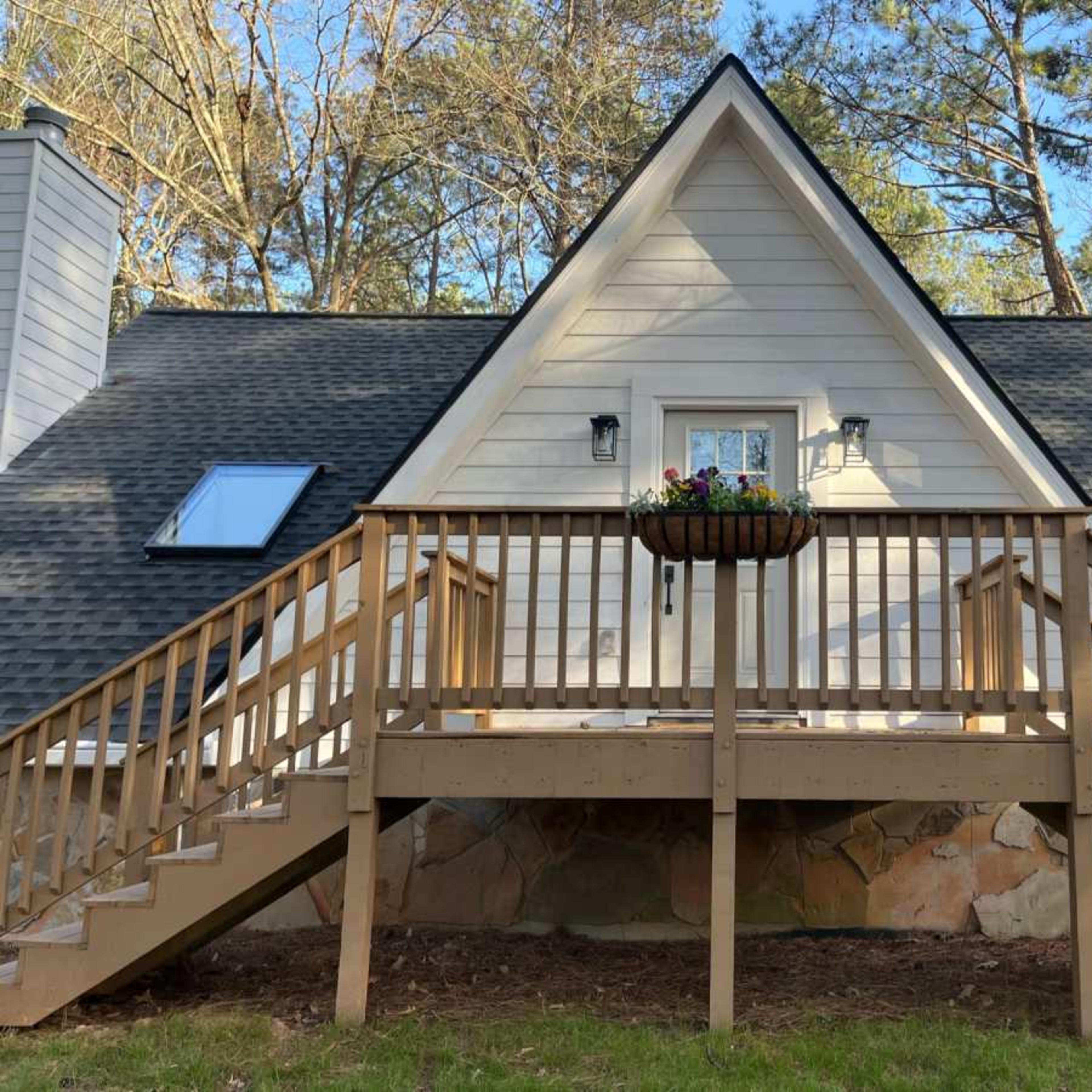 A triangular-roofed house with a wooden deck and stairs in a forested area.