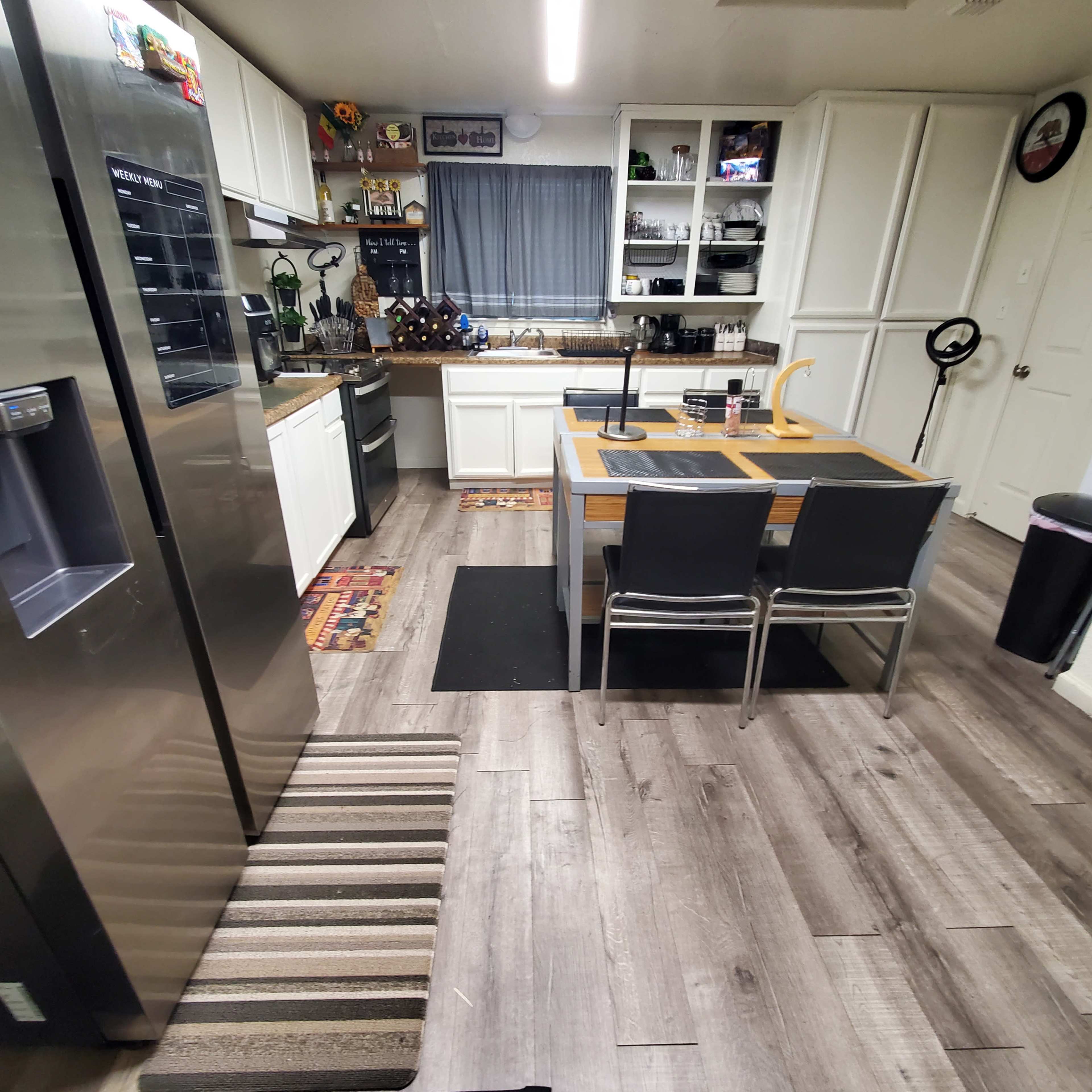 A modern kitchen with a stainless steel refrigerator, a wooden dining table set with chairs, and gray laminate flooring.