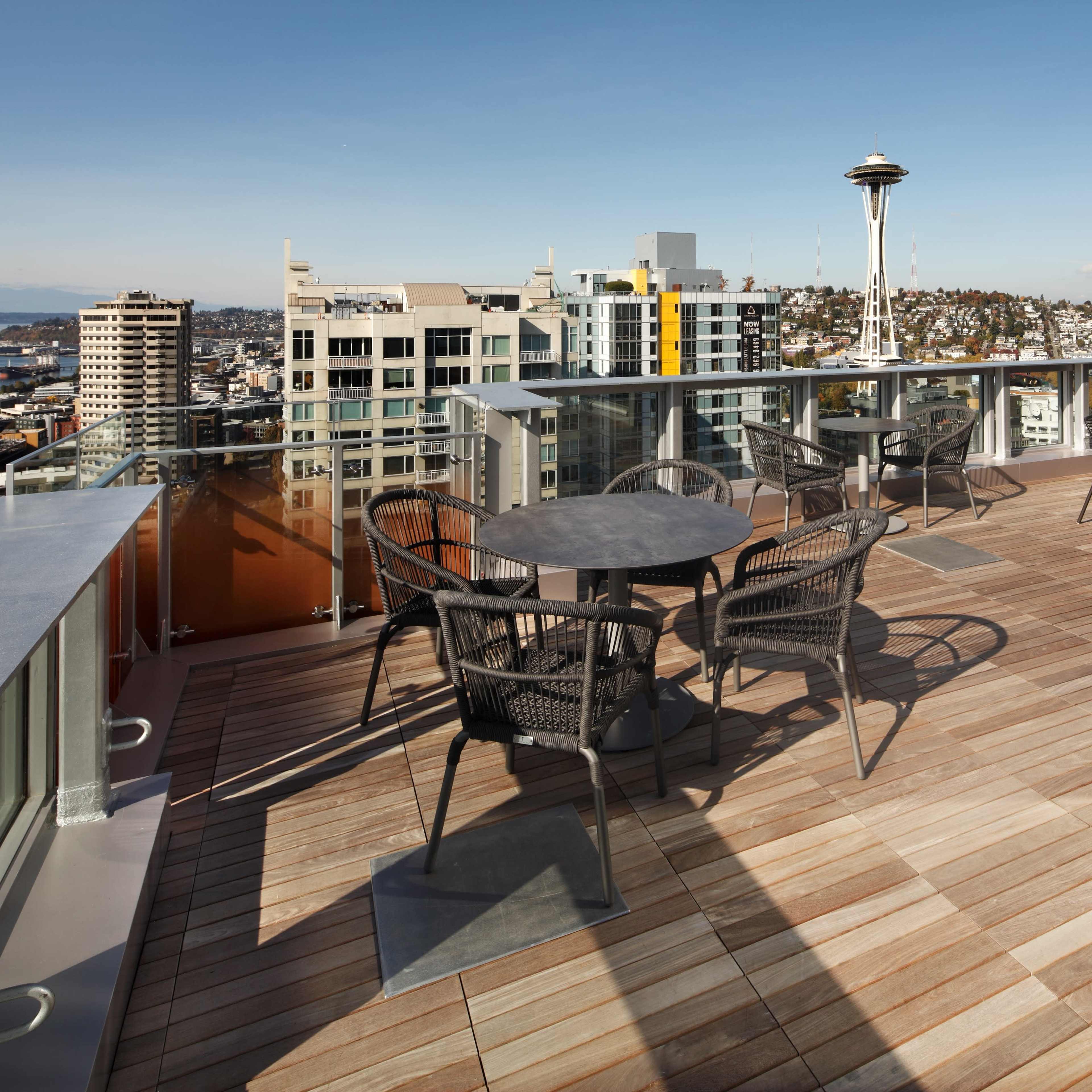 A rooftop terrace with several round tables and chairs overlooking a cityscape, including the Space Needle in the distance.