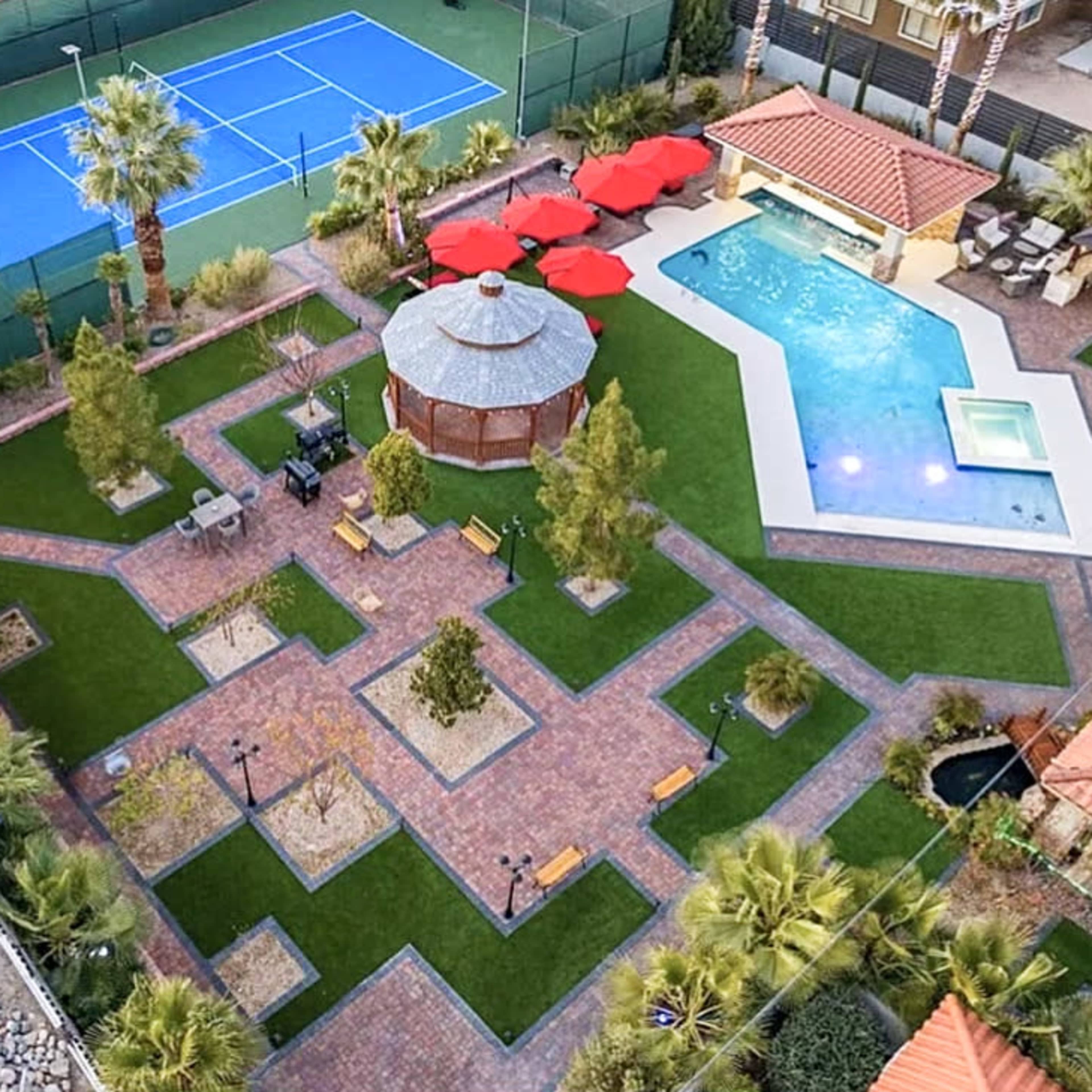 A landscaped outdoor area featuring a swimming pool, a gazebo, tennis courts, and surrounding greenery with pathways and seating areas.