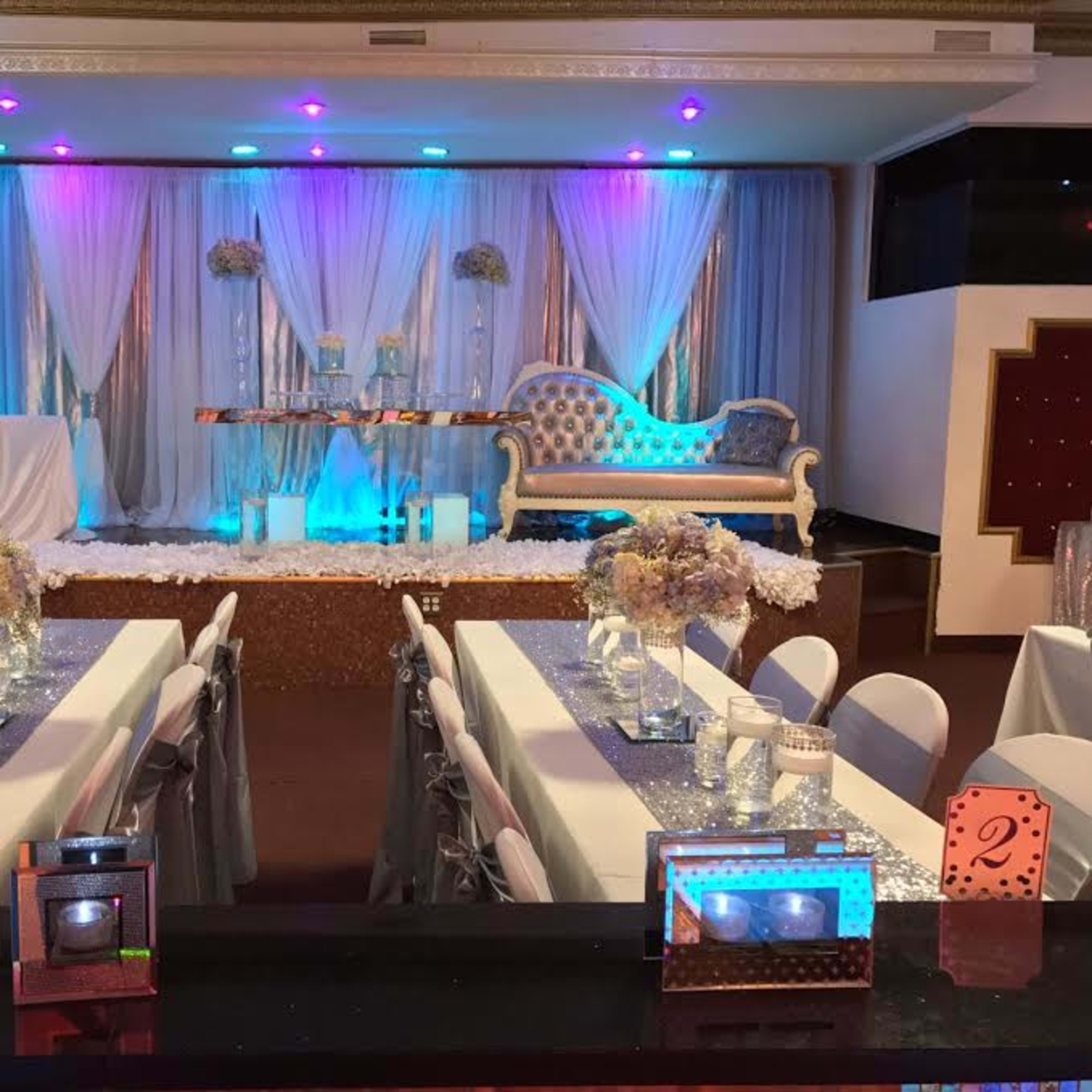 A banquet hall decorated with white and blue drapes, a stylized stage with floral arrangements, and tables set with elegant centerpieces.