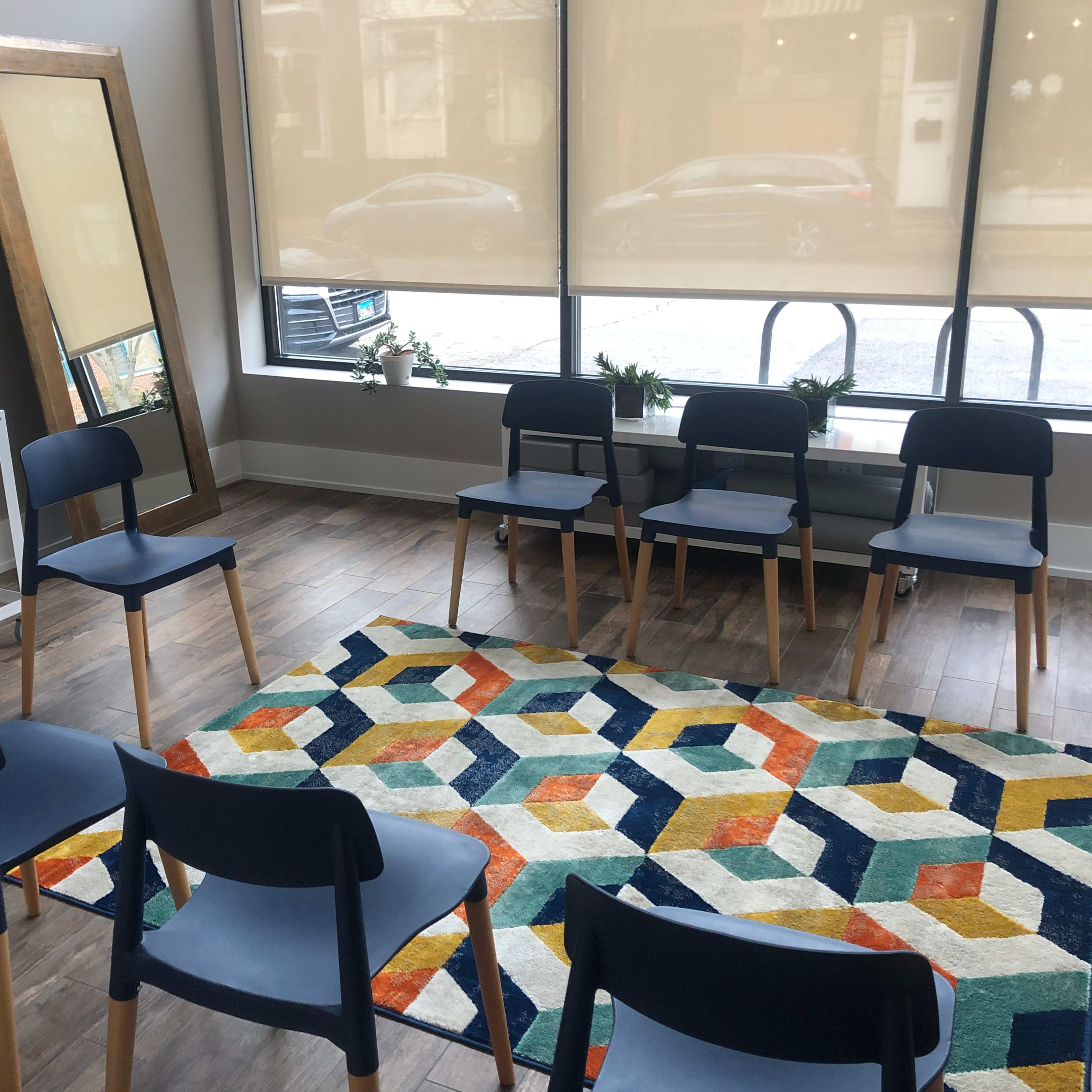 A circle of blue chairs surrounds a patterned rug in a brightly lit room with large windows and a mirror.