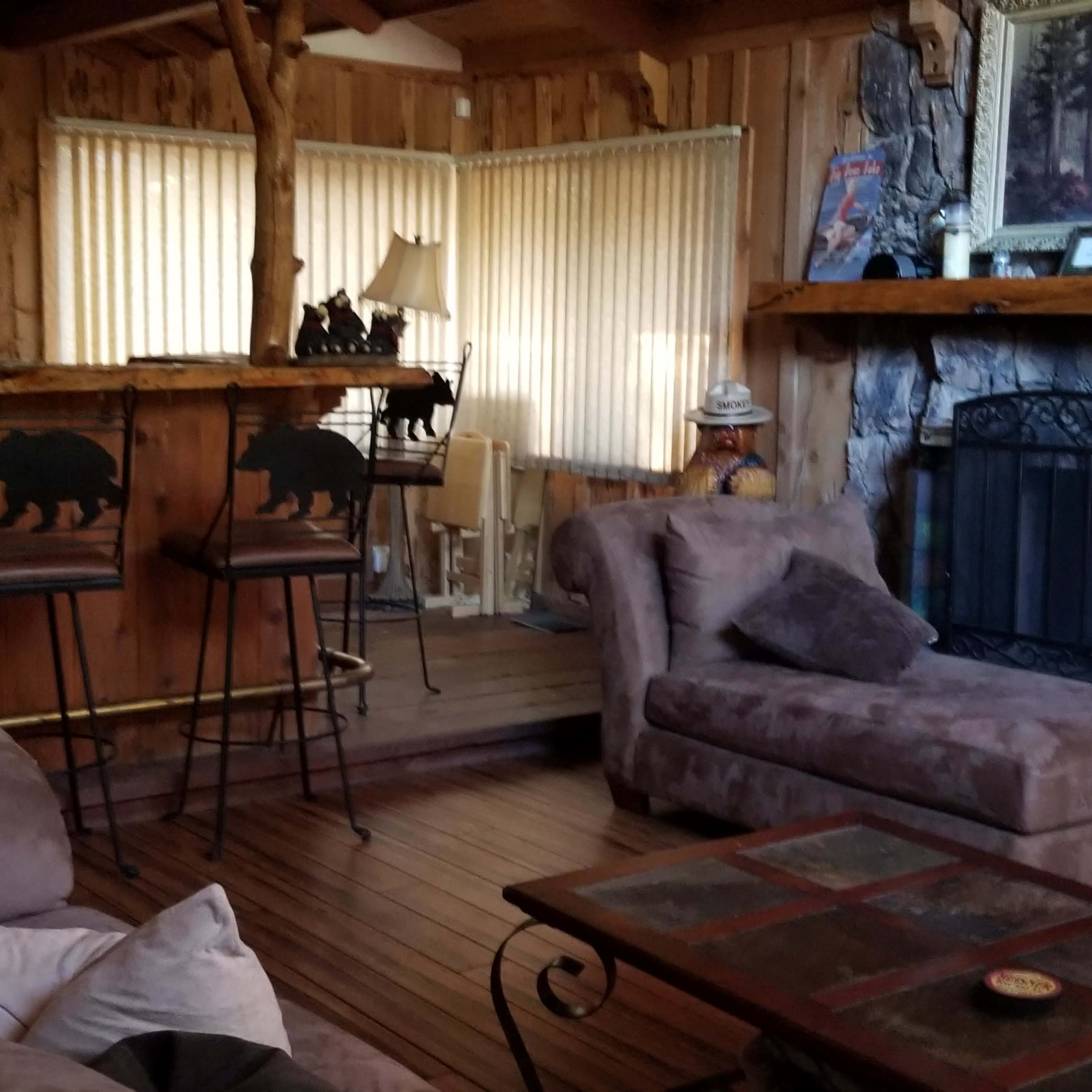 A cozy living room features a stone fireplace, wooden paneling, a large brown sofa, and a bar area with bear-themed stools.