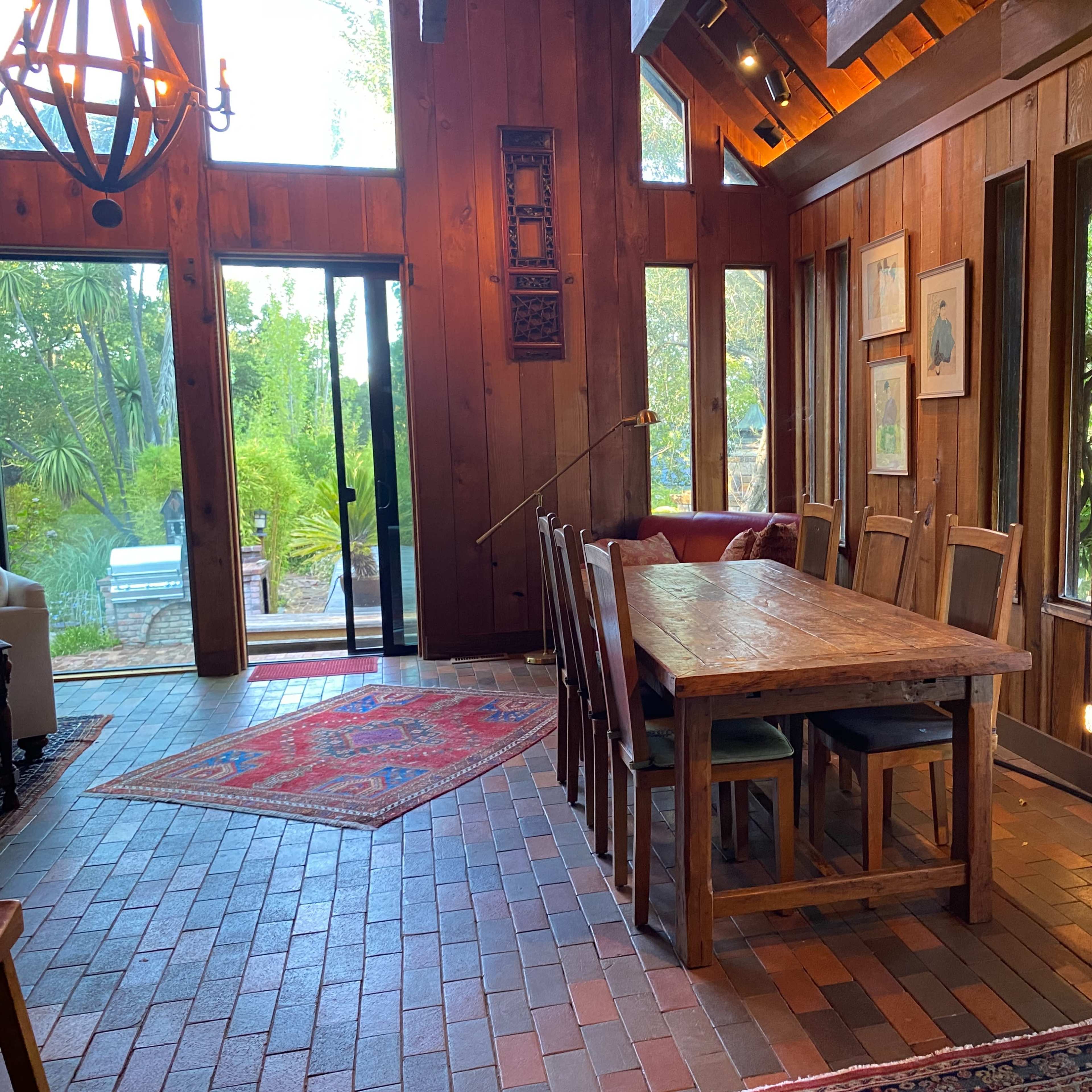 A wooden dining area with a large table and chairs, illuminated by natural light from surrounding windows, next to a patio door leading to an outdoor space.