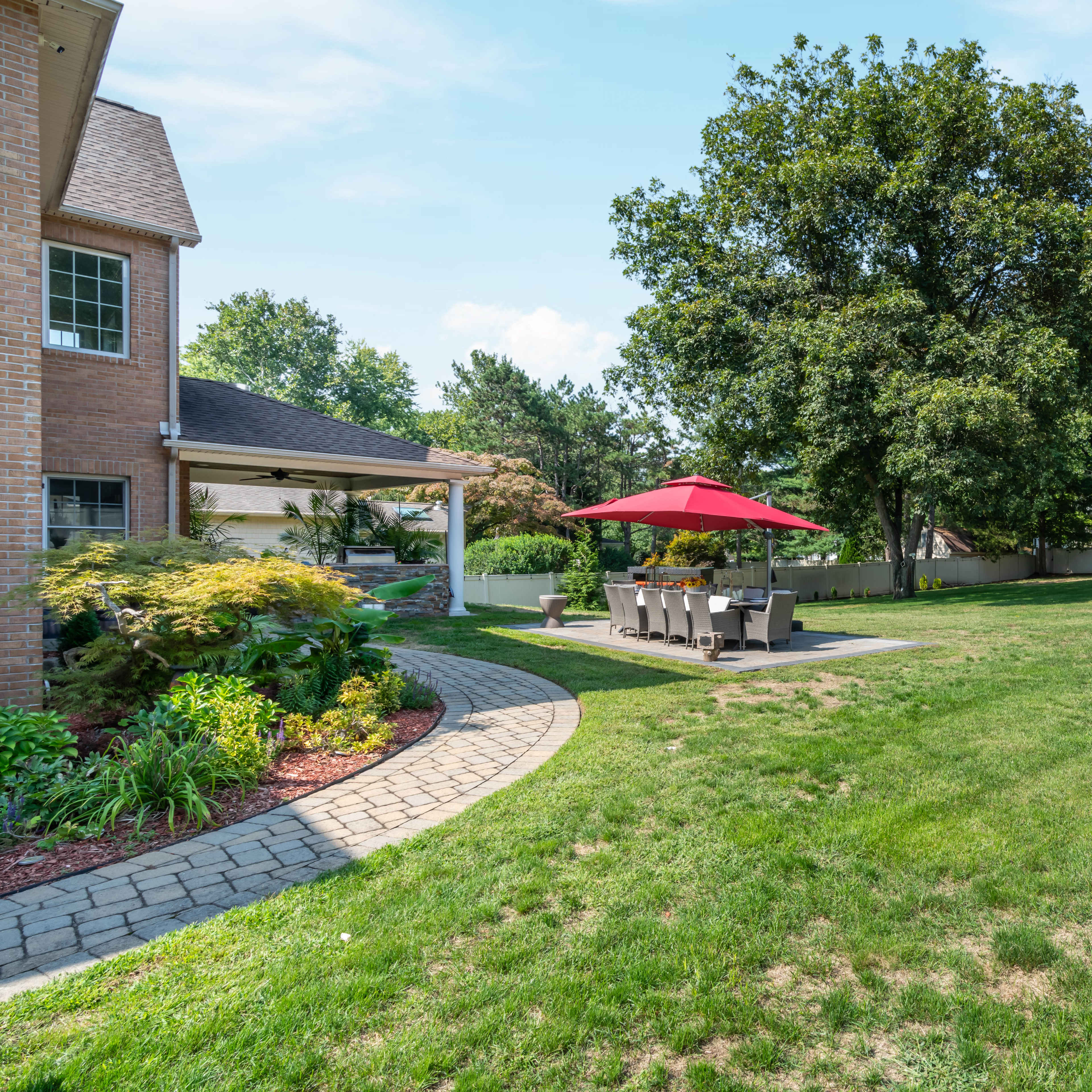 A spacious backyard features a stone pathway leading to a patio with an umbrella-covered table, surrounded by grass and trees.