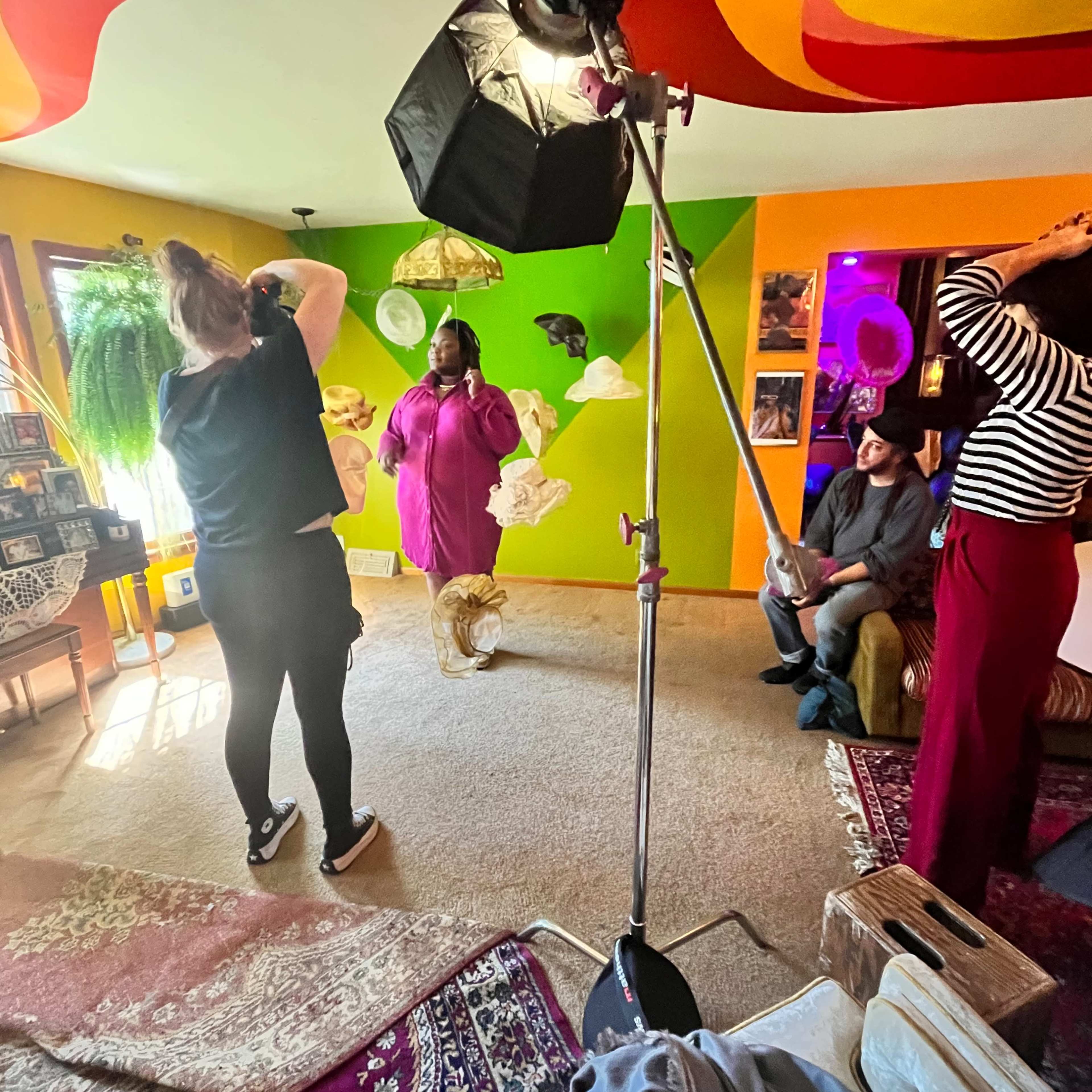 A person is photographing a model sitting in a brightly colored, plants-filled living room with eclectic decor.