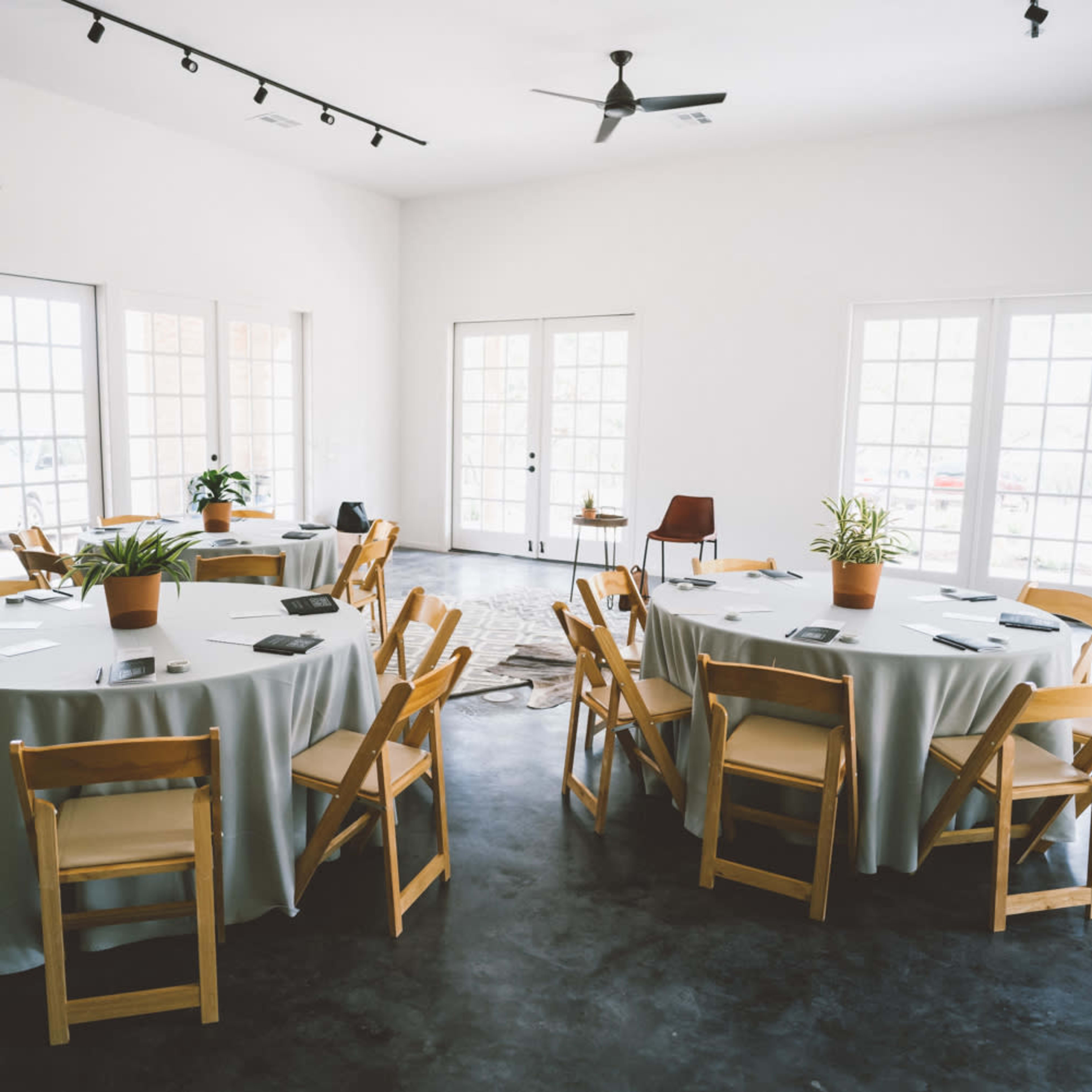 A bright room features multiple round tables with chairs, surrounded by large windows and a concrete floor.