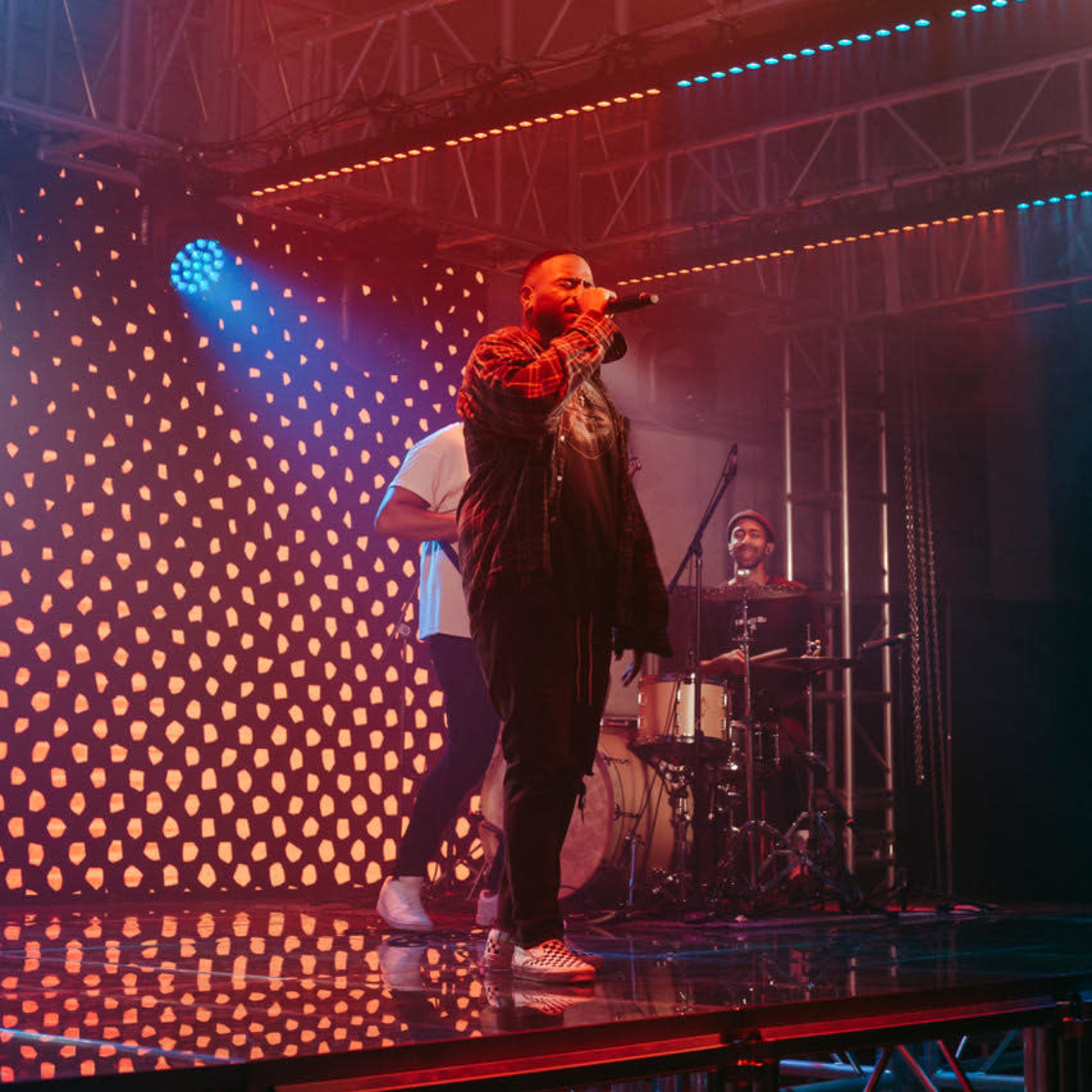 A musician sings into a microphone on stage while a drummer plays in the background under vibrant lighting.