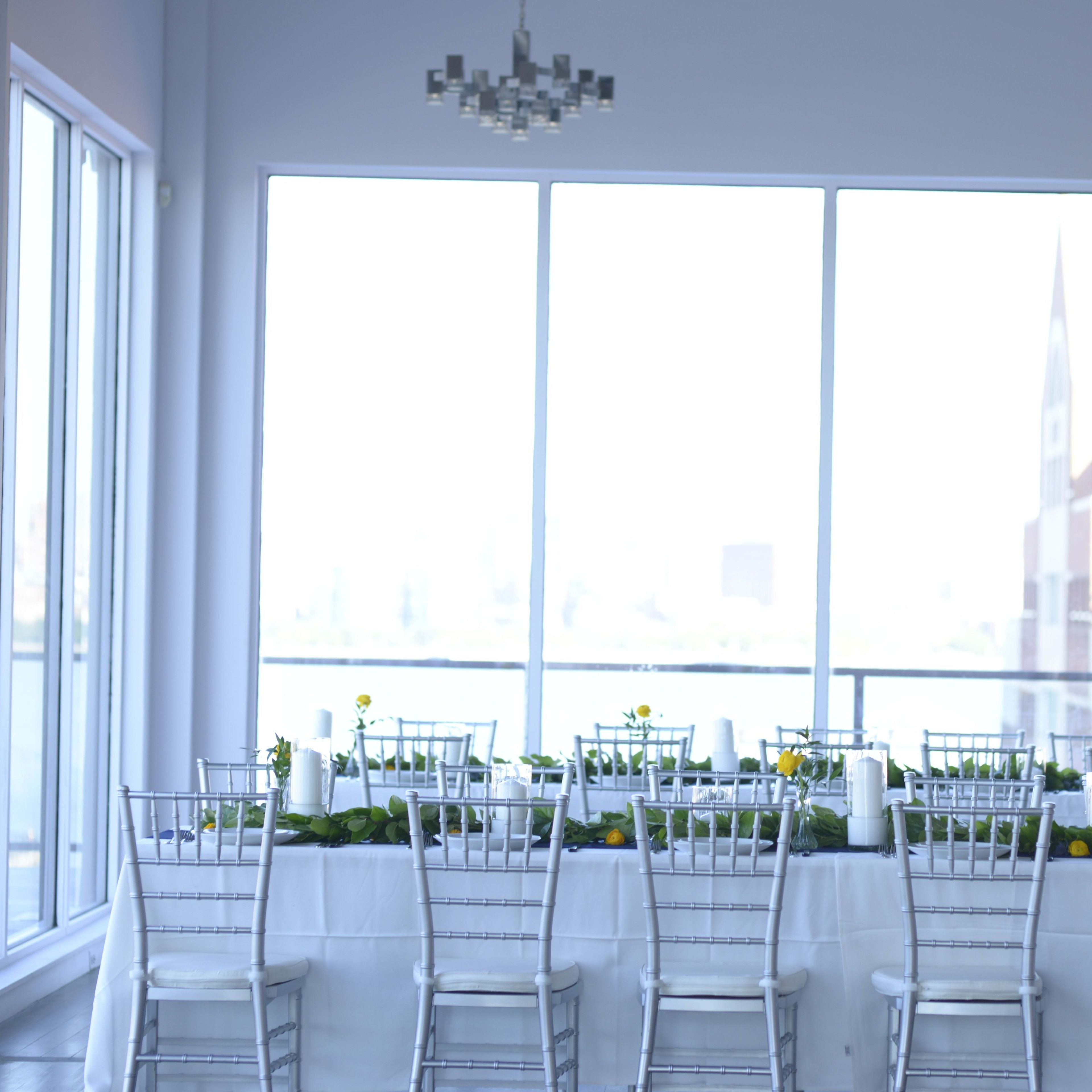 A banquet hall features a long, elegantly set table adorned with greenery and candles, positioned near large windows that provide ample natural light.