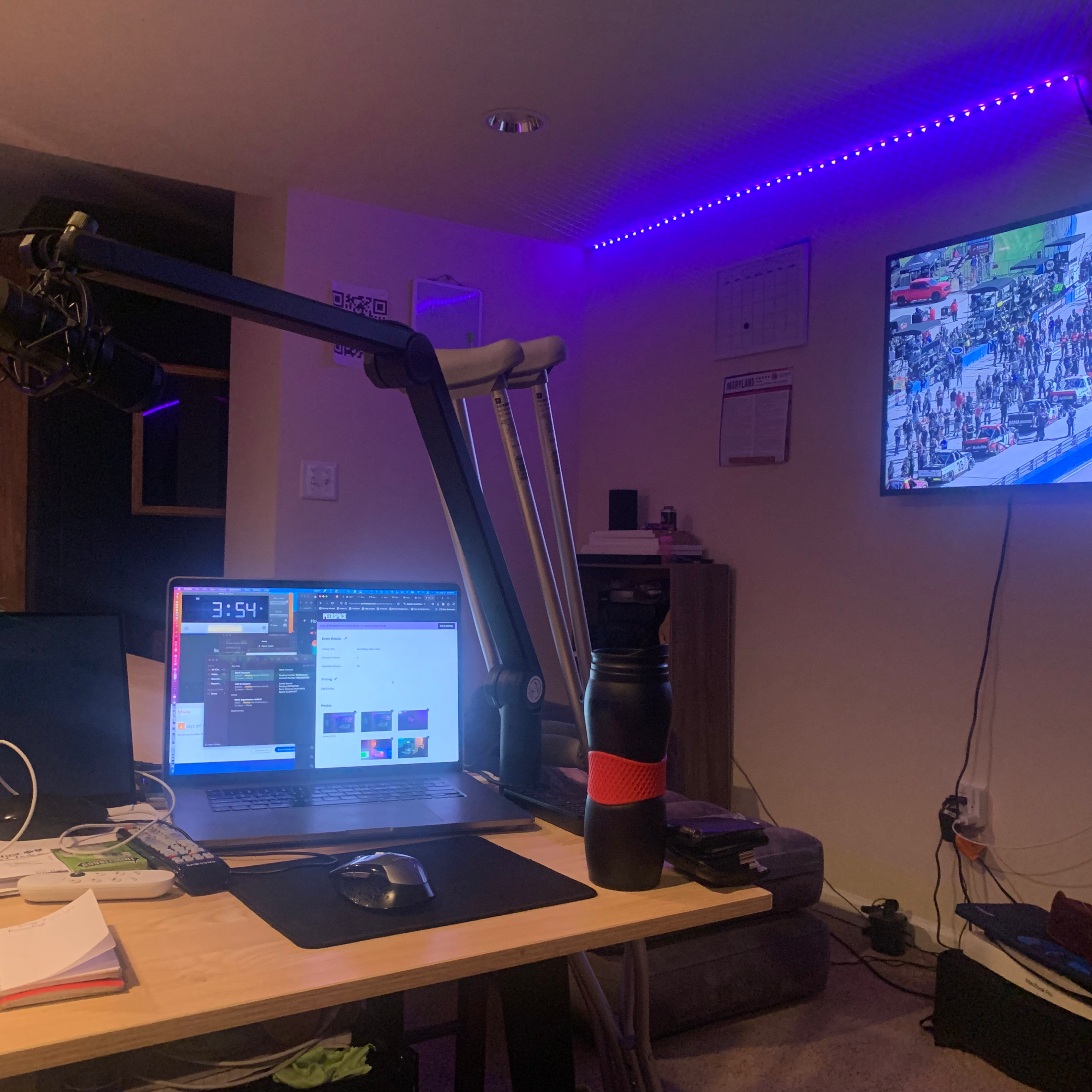 A home workspace with a computer, microphone on an adjustable arm, a water bottle, and a television displaying a sports event, all set in a dimly lit room with LED ambient lighting.