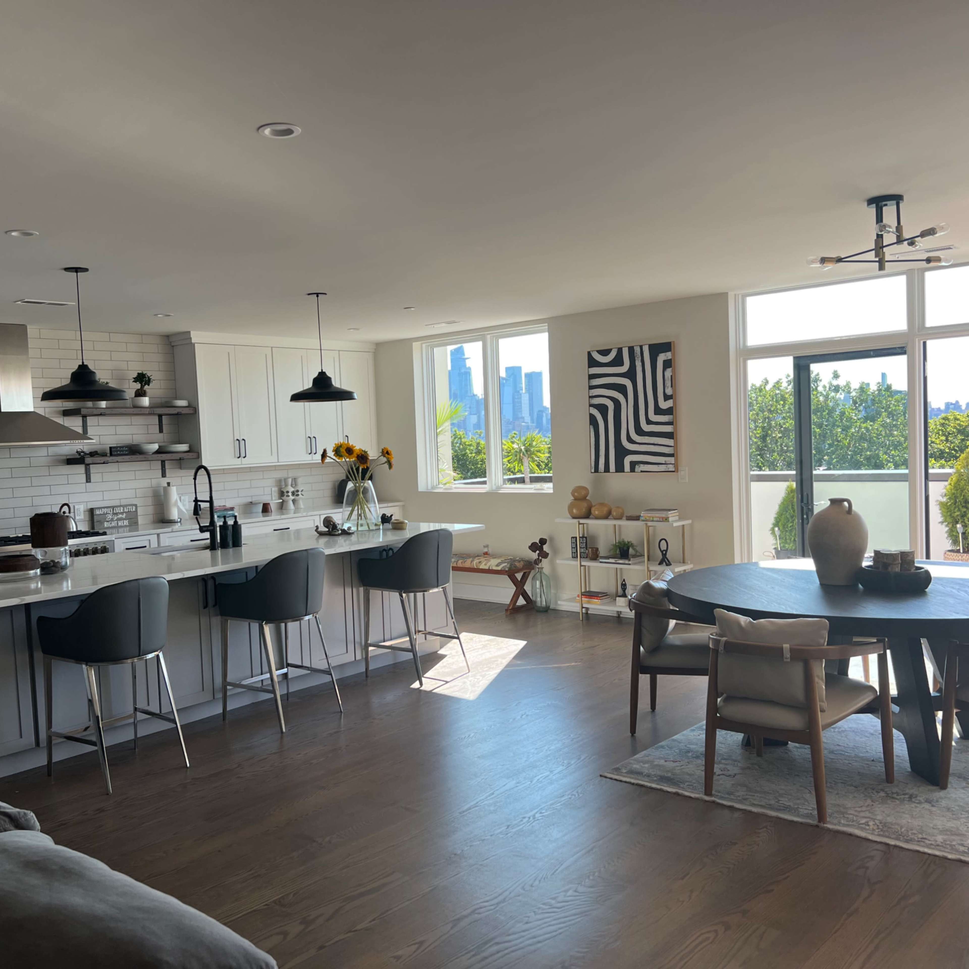 A modern kitchen and dining area with a large island, seating for four, and a panoramic view of a city skyline through large windows.