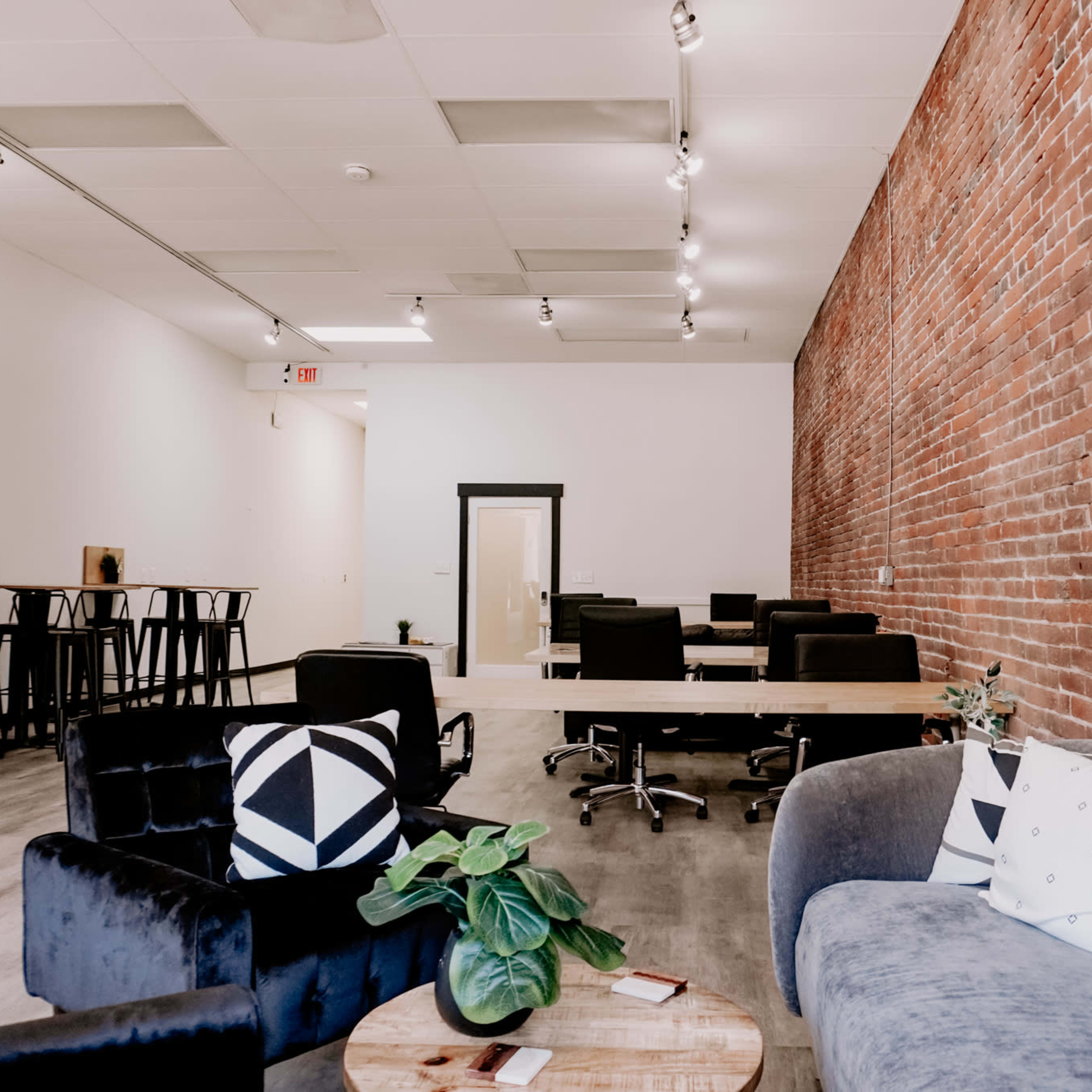 A modern office space featuring a combination of a brick wall, workstations with chairs, and a lounge area with couches and plants.