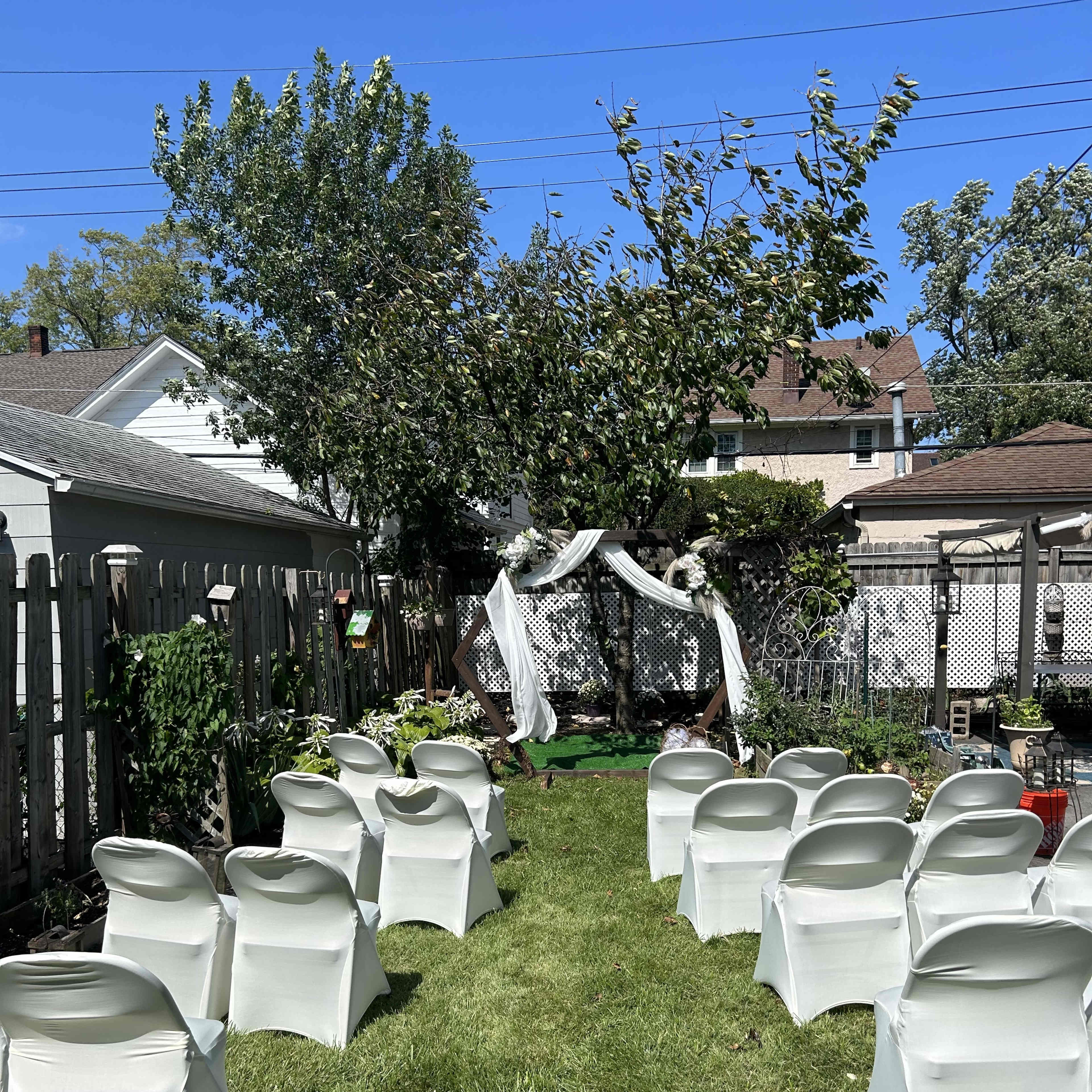 A garden is set up for a ceremony with white chairs lined up on a grassy area facing a decorated arch under a tree.