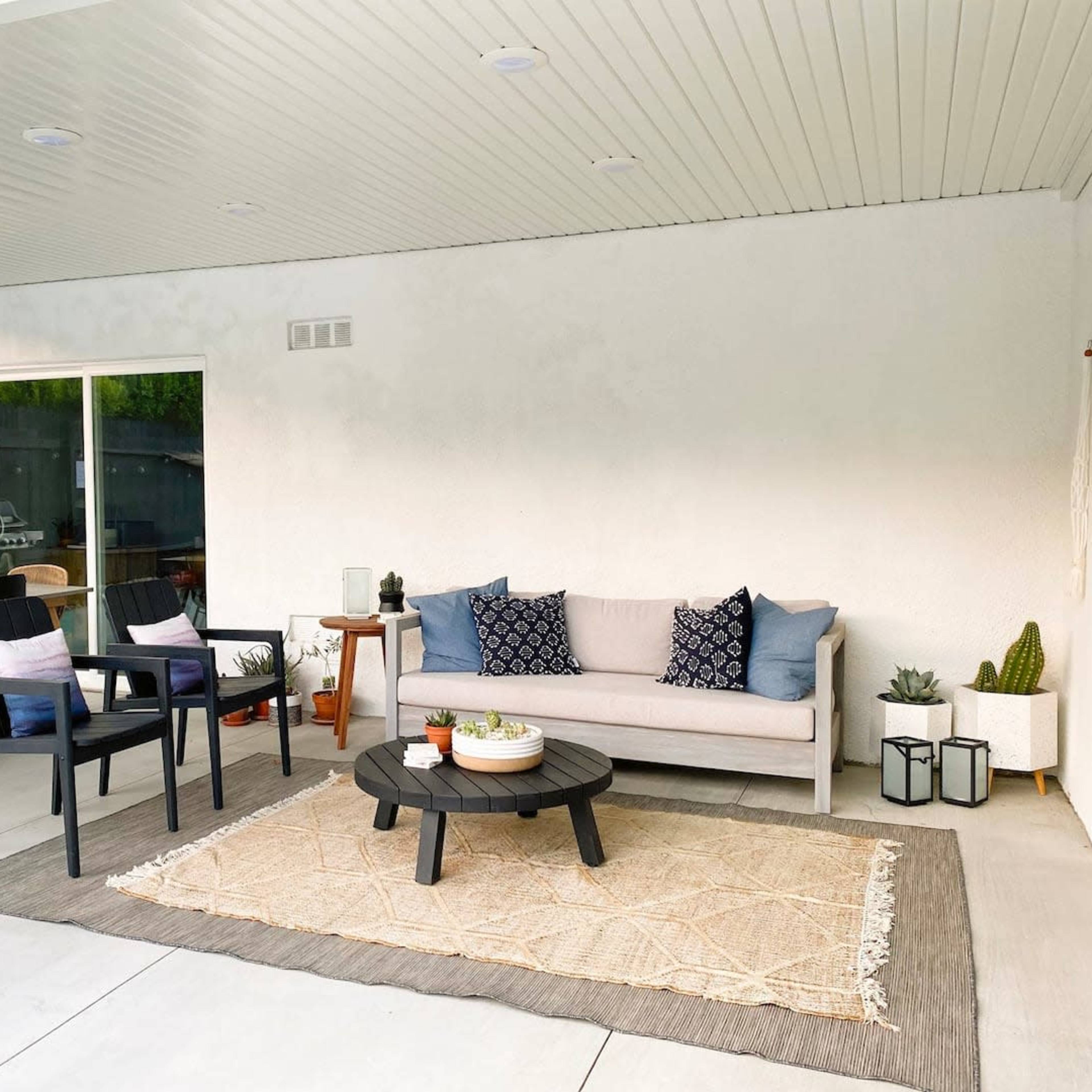 A covered outdoor seating area featuring a couch, two chairs, a round coffee table, and decorative plants on a light-colored rug.