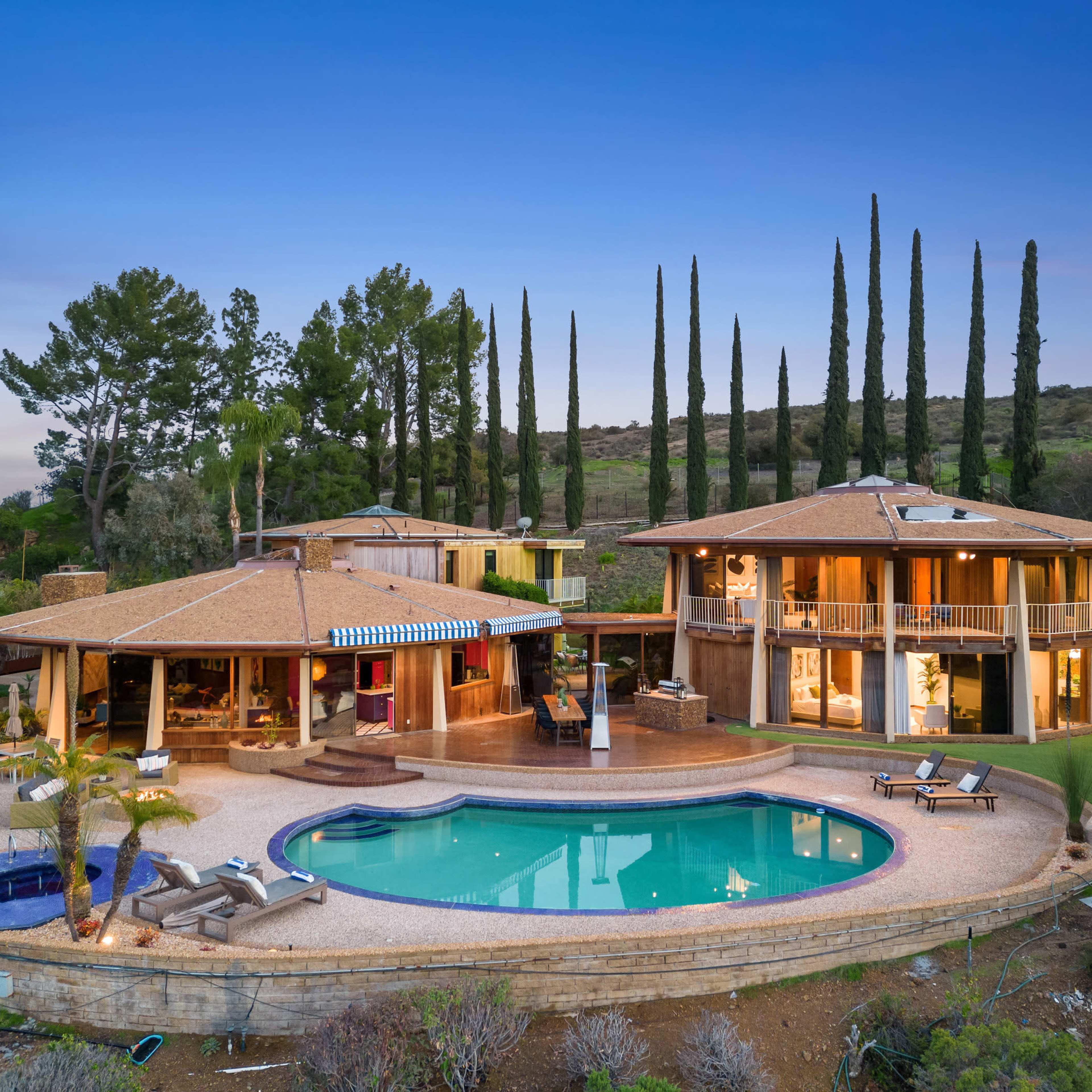 A modern two-story house with a rounded pool and landscaped yard is set against a backdrop of tall cypress trees and rolling hills.