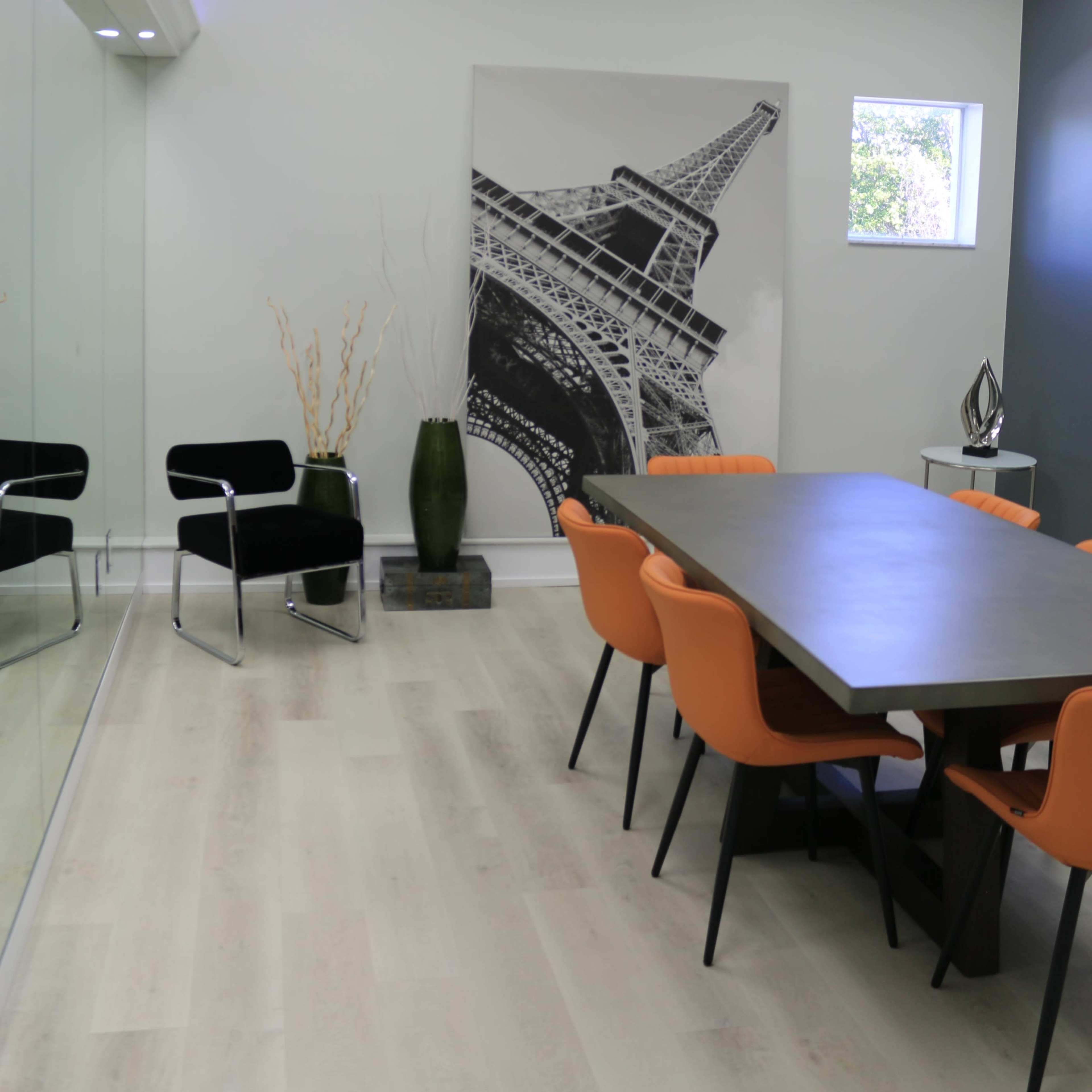 A modern conference room features a large gray table surrounded by orange chairs, a black-and-white Eiffel Tower poster on the wall, and a glass partition.