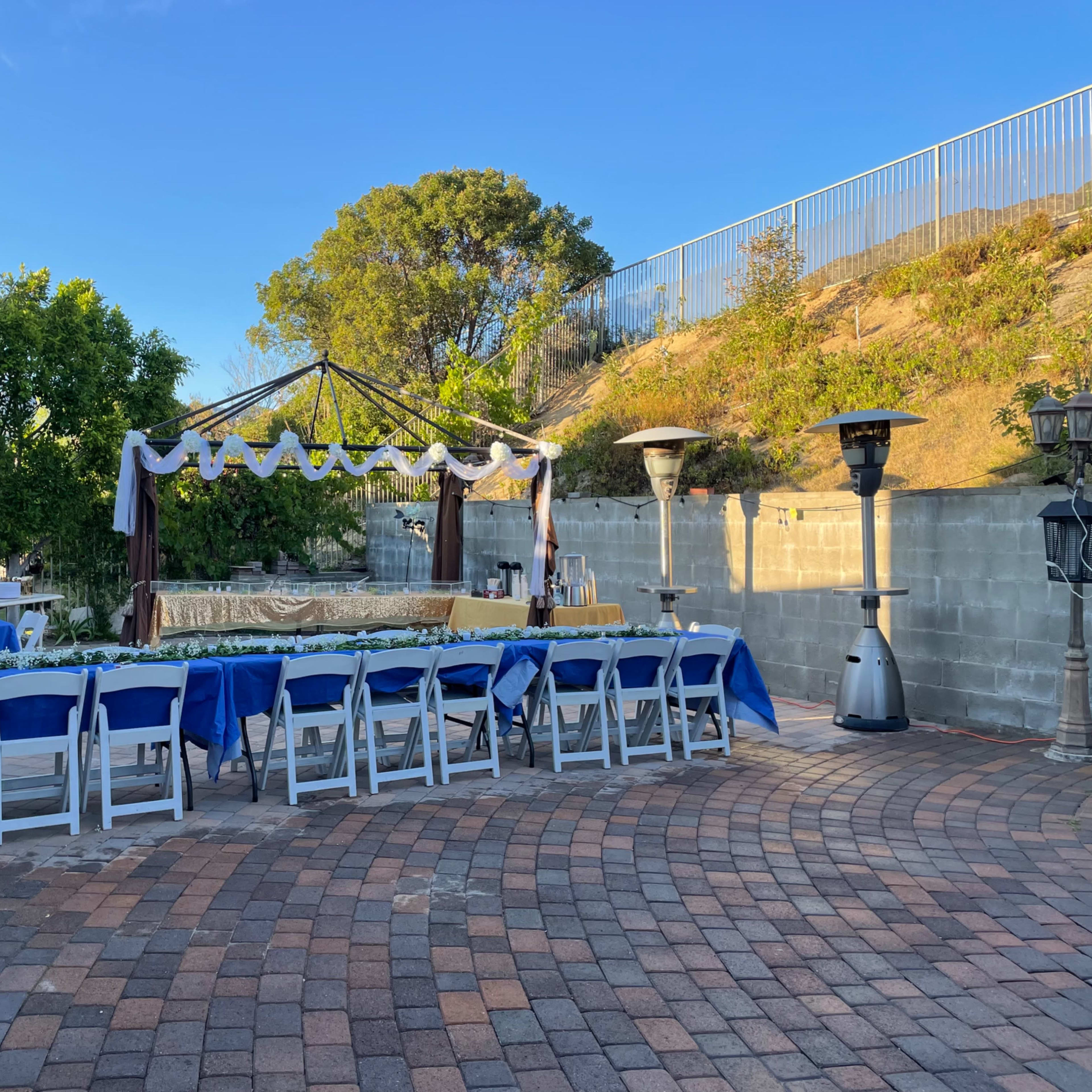 A set-up for an outdoor event features rows of blue-covered tables and a decorated canopy in a garden area.