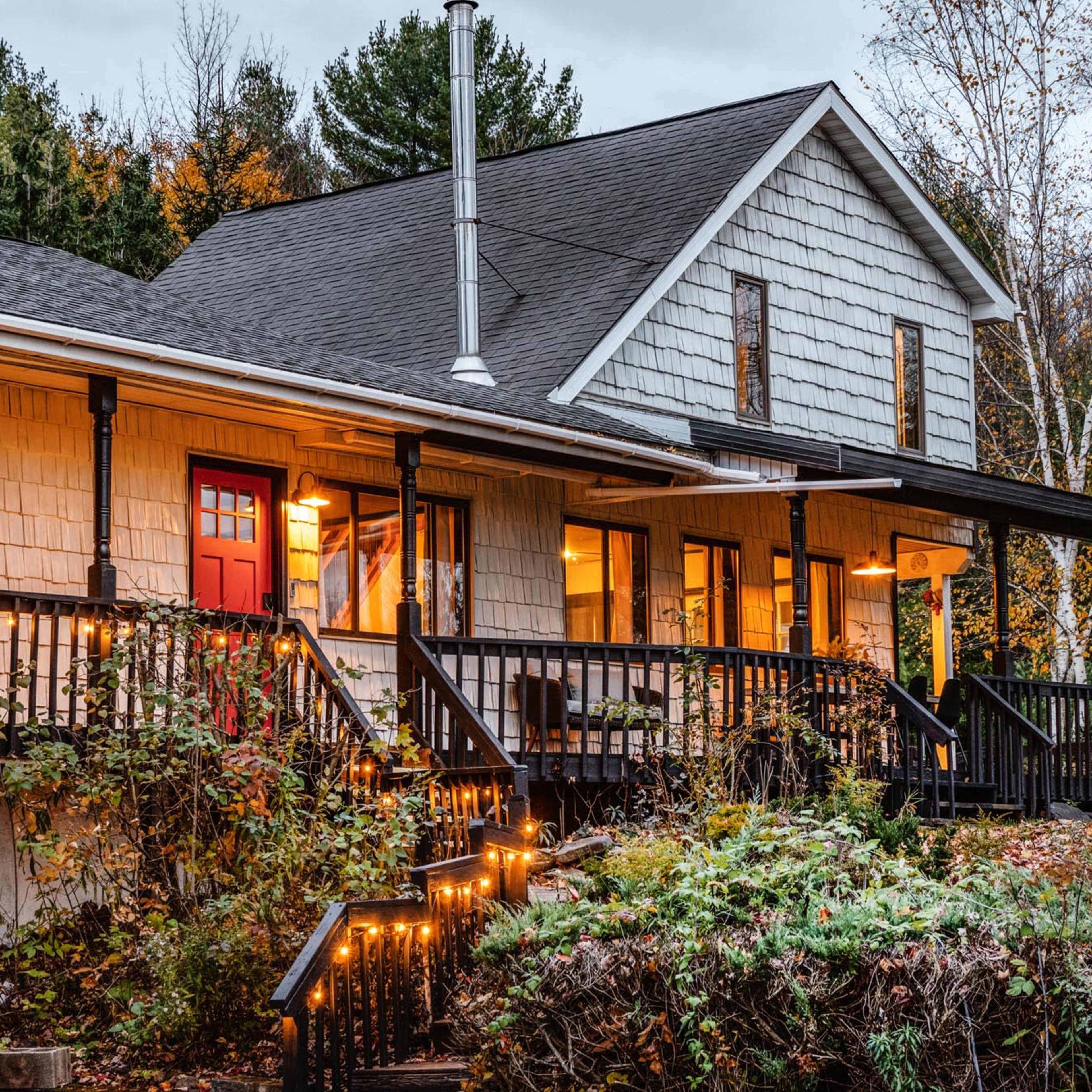 A house with a shingled facade and red door is surrounded by autumn foliage and features a front porch with lights and wooden railings.