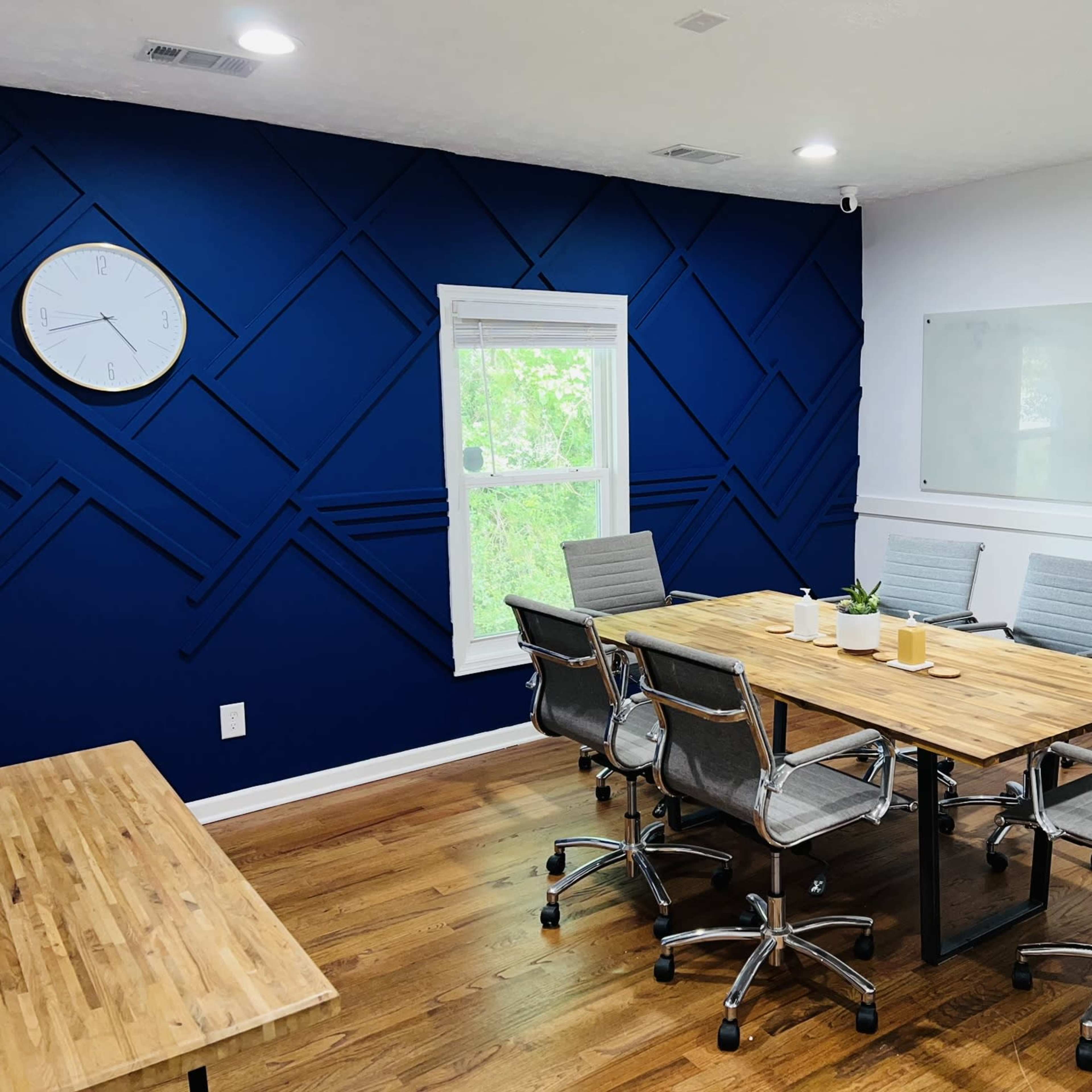 A modern conference room features a wooden table surrounded by ergonomic chairs, with a large clock on the wall and a blue accent wall behind it.