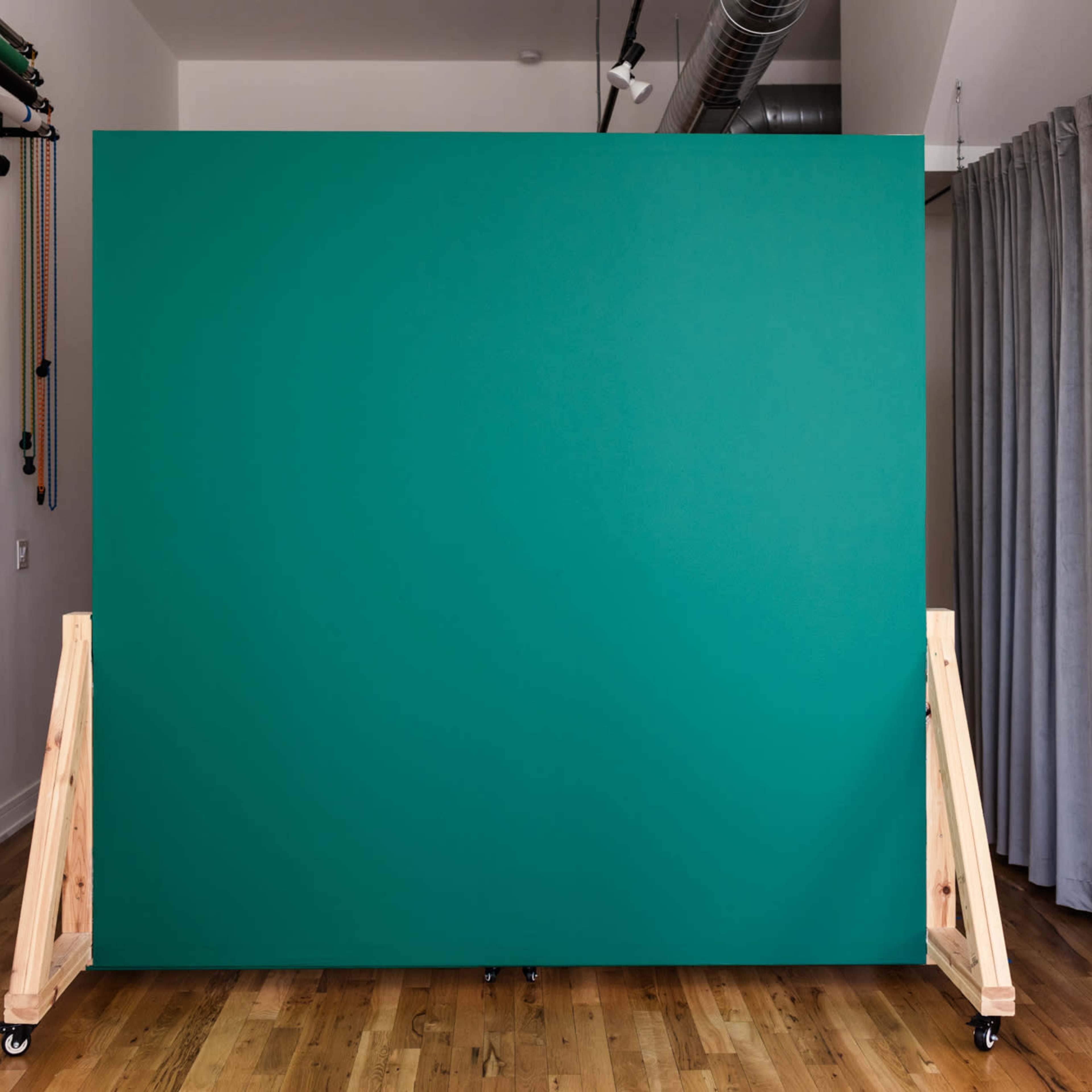 A green backdrop mounted on wooden stands in a room with light-colored wooden flooring and gray curtains.