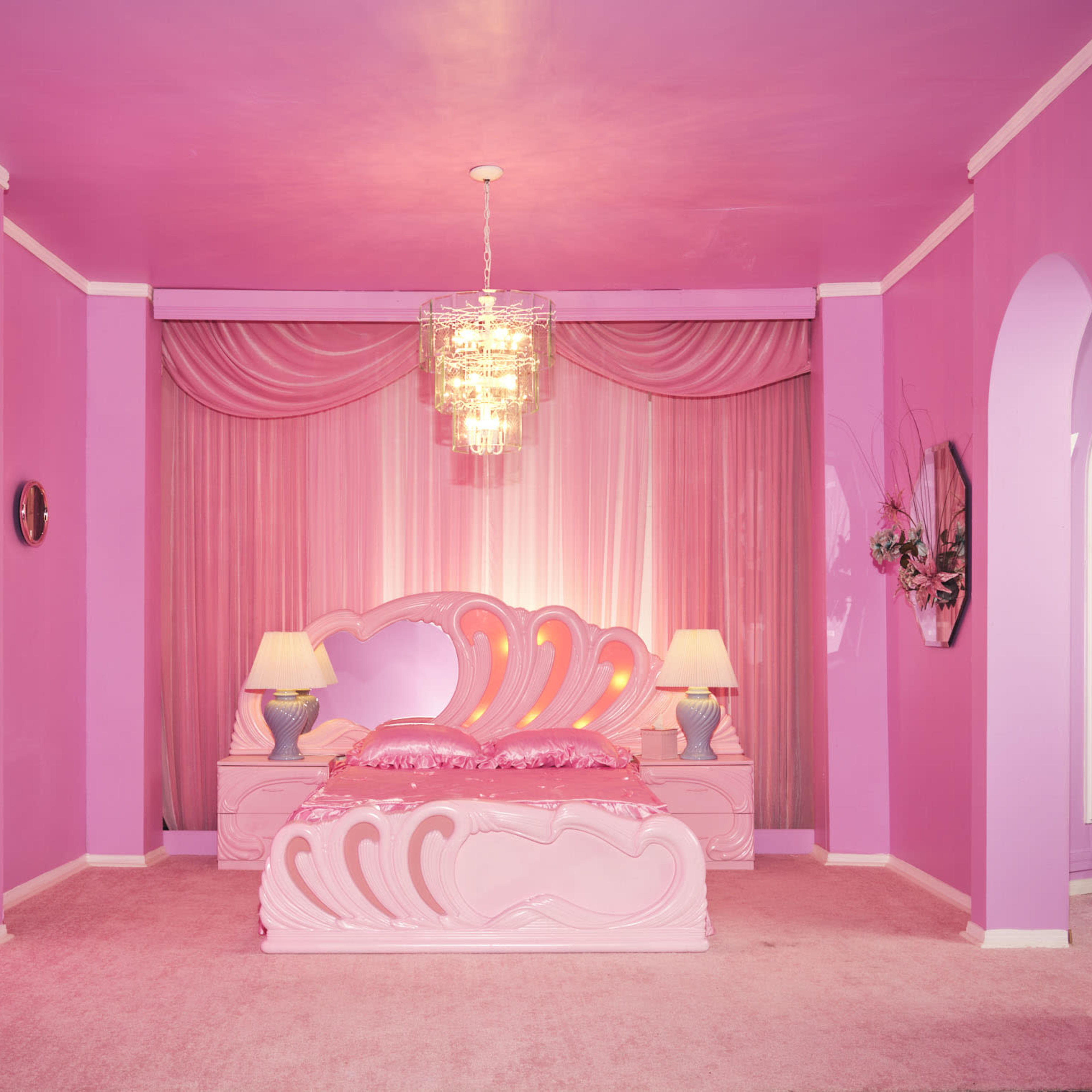 A brightly colored pink bedroom features a heart-shaped bed, matching lamps, a chandelier, and archways leading to other areas.