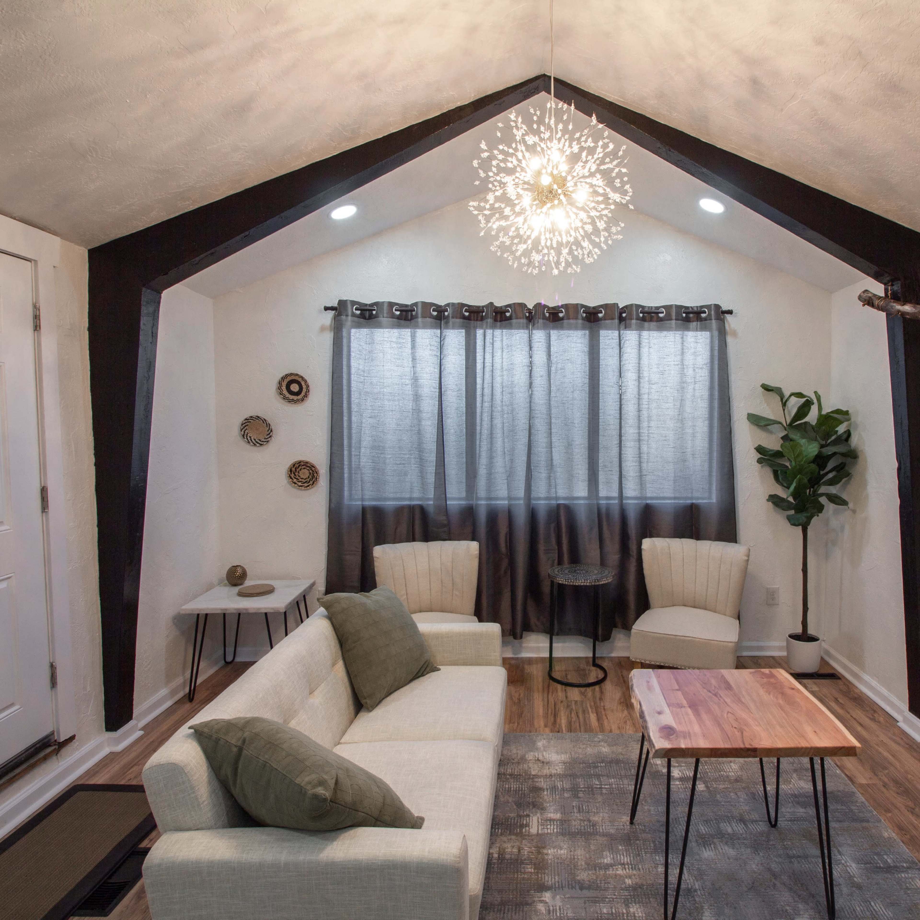 A modern living room features a sofa, two armchairs, a wooden coffee table, a decorative plant, and a wall-mounted TV, all illuminated by a distinctive pendant light and natural light from the window.